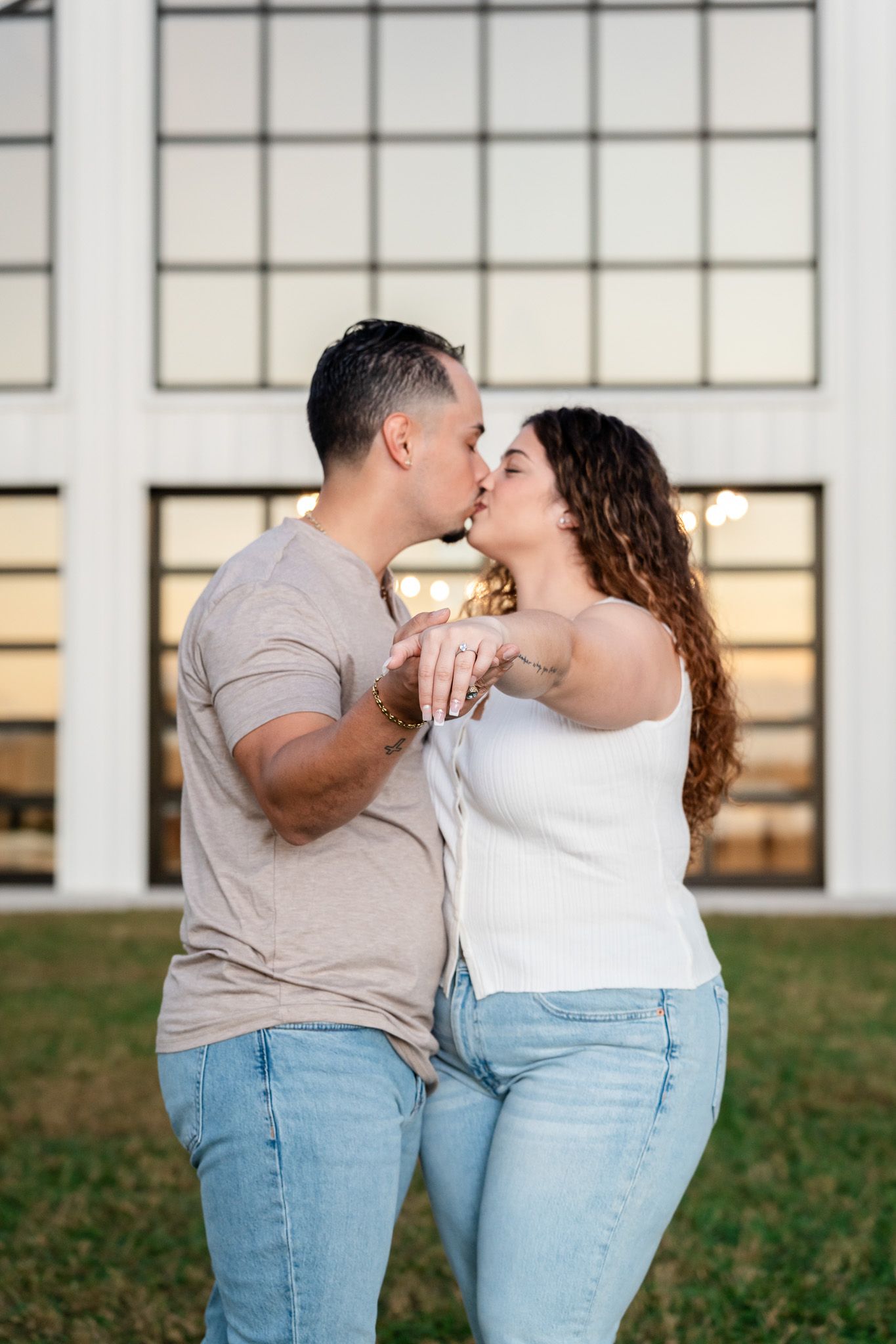 Engaged couple shares a kiss while showing off their engagement ring outdoors.