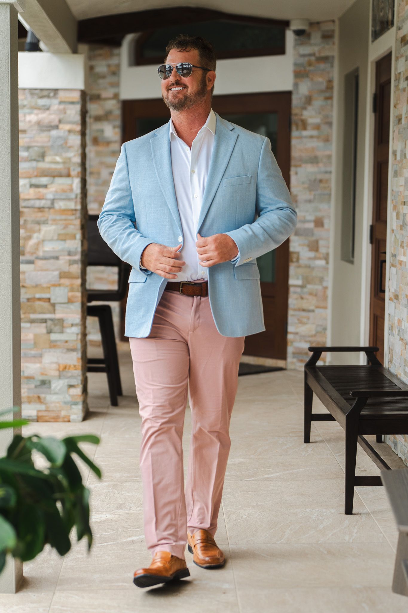 Smiling man in light blue blazer, pink pants, and sunglasses walking along a stone-columned outdoor corridor.
