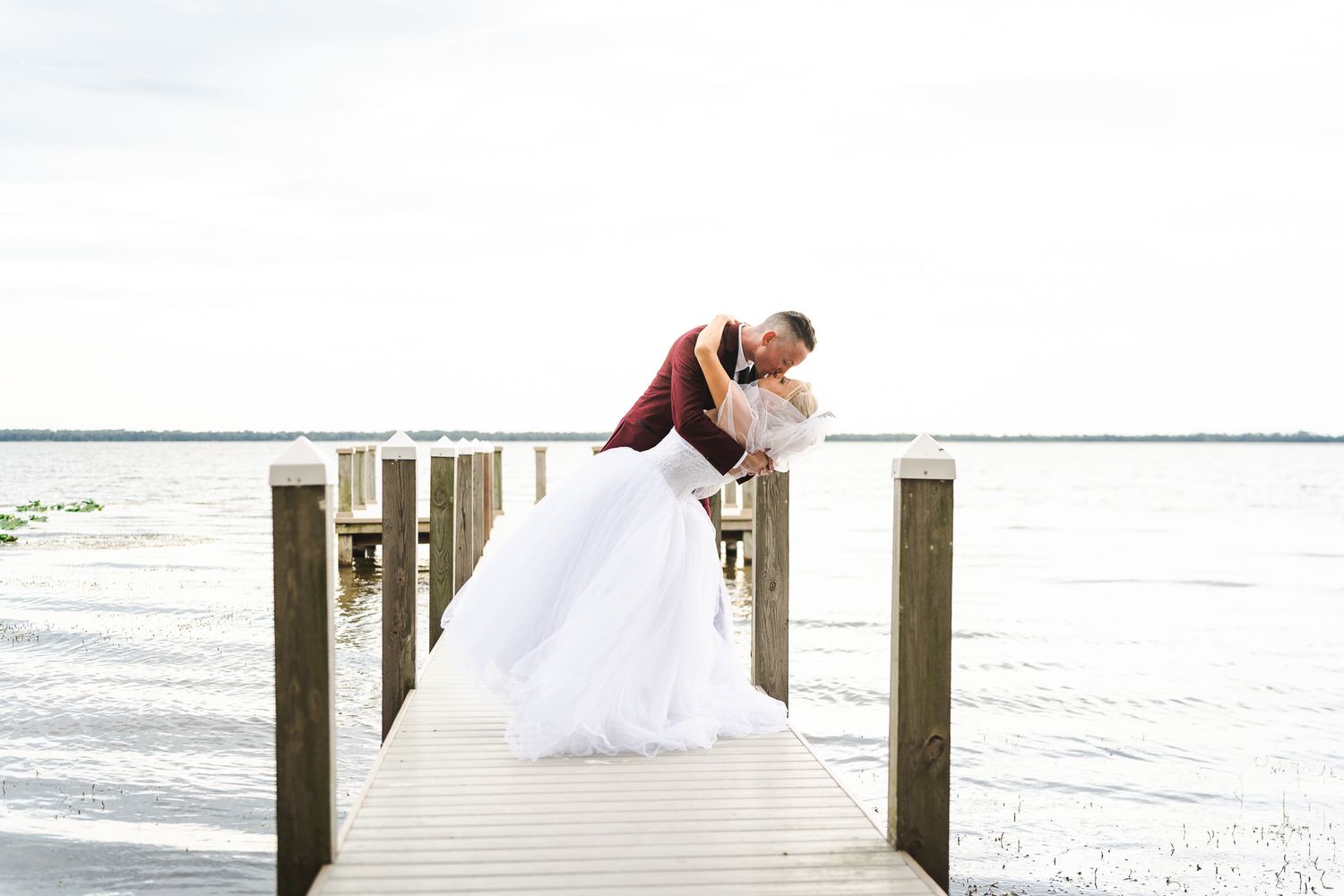 Groom dipping bride in romantic pose at end of wooden dock over lake.