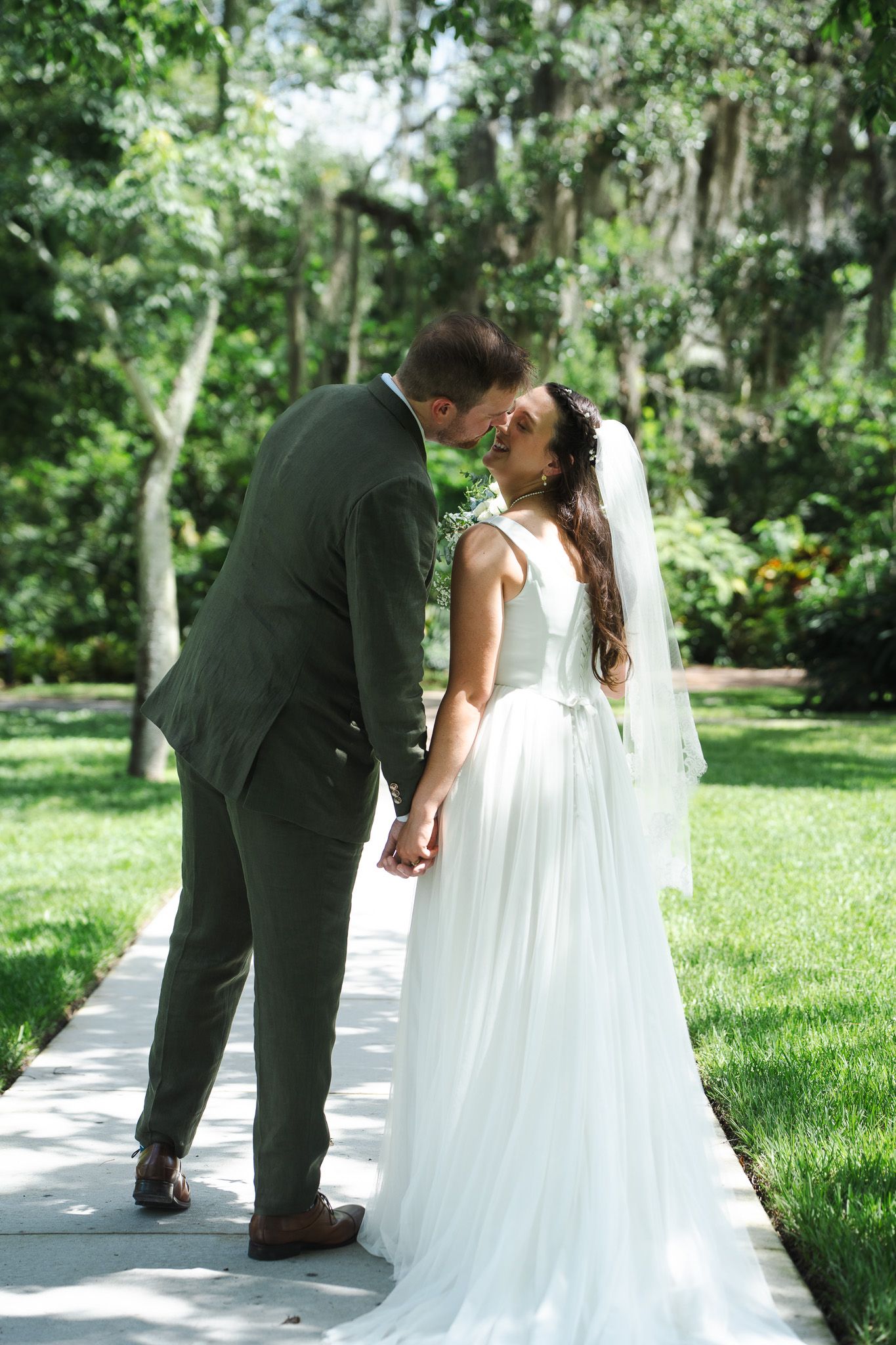 Bride in white tulle gown and groom in olive suit share a romantic kiss on a garden pathway.