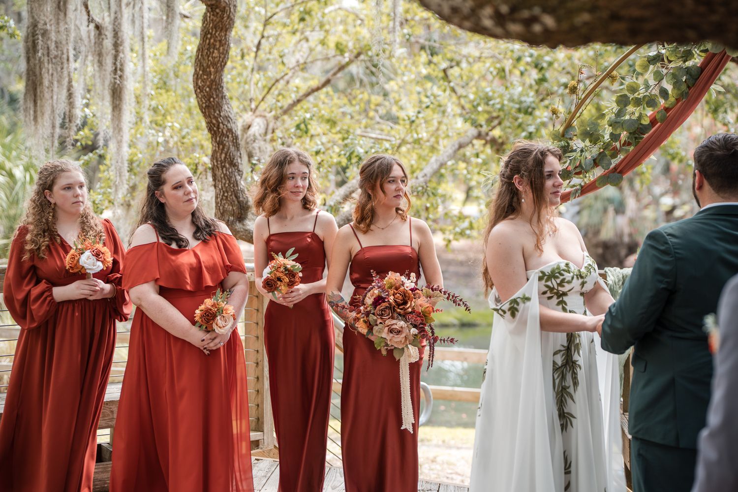 Five bridesmaids in rust-colored dresses holding bouquets stand beside bride at outdoor wedding ceremony.