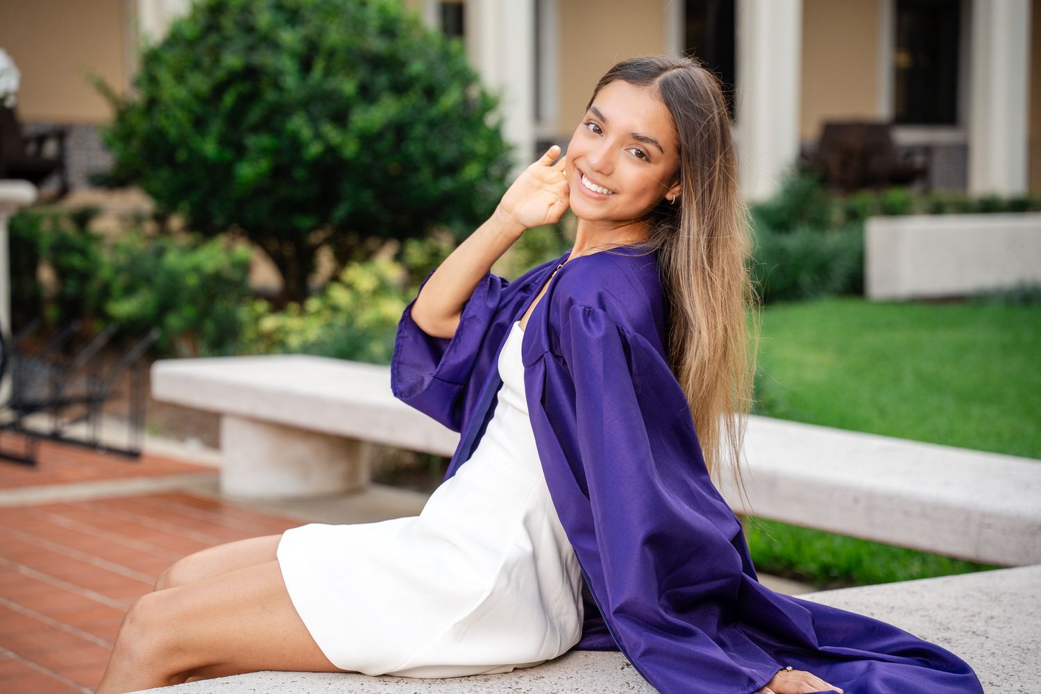 Young woman in purple graduation gown and white dress sitting outdoors, smiling at camera on a stone bench.