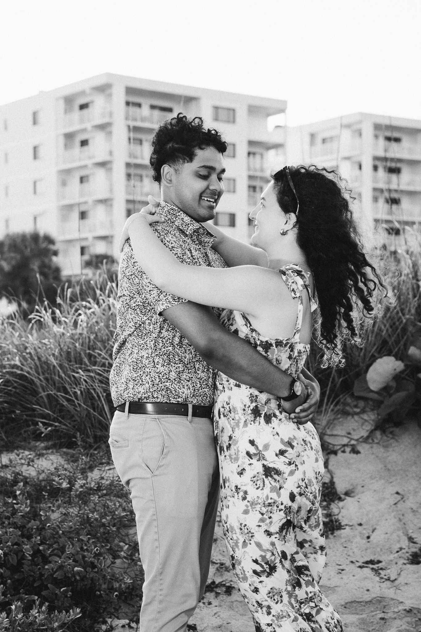 Black and white photo of a couple embracing and smiling at each other on a sandy beach with buildings in background.