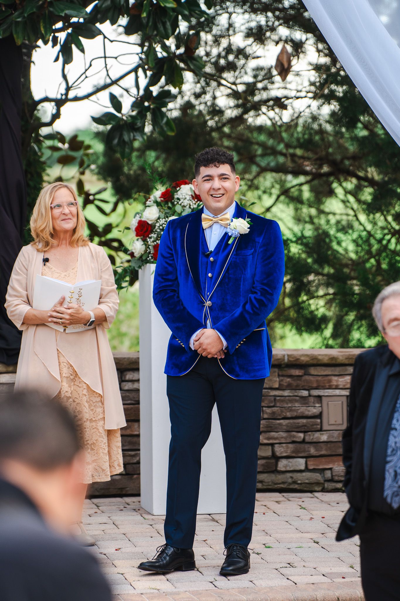 Groom in royal blue velvet tuxedo smiling at altar during outdoor wedding ceremony.