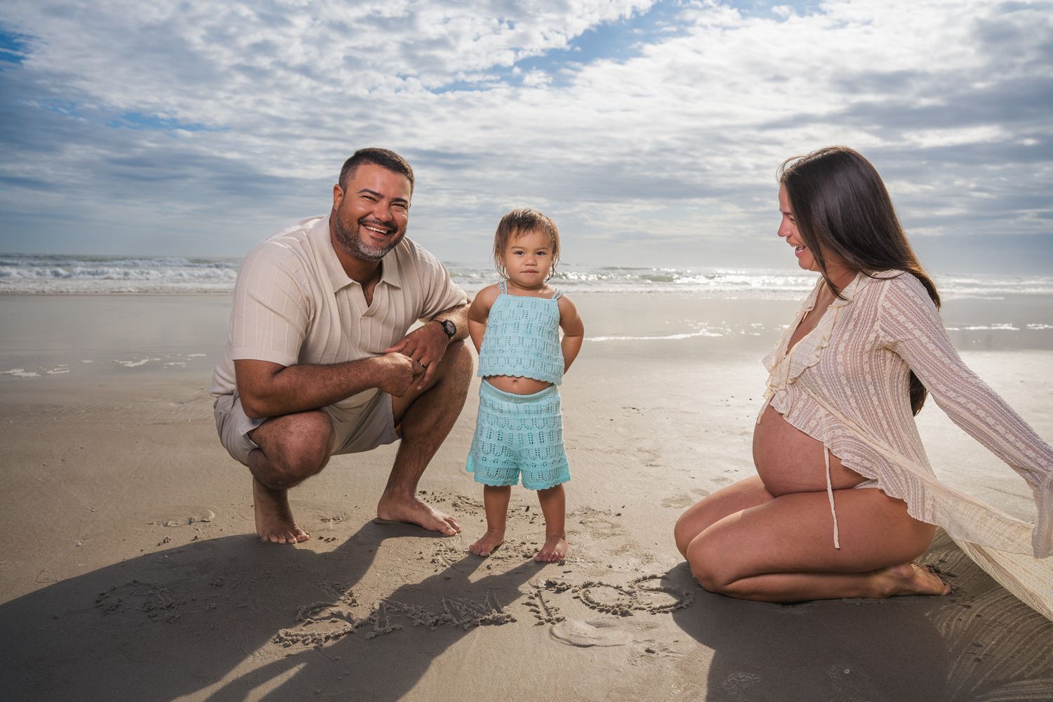 Family maternity photo with father, toddler, and pregnant mother on sandy beach.