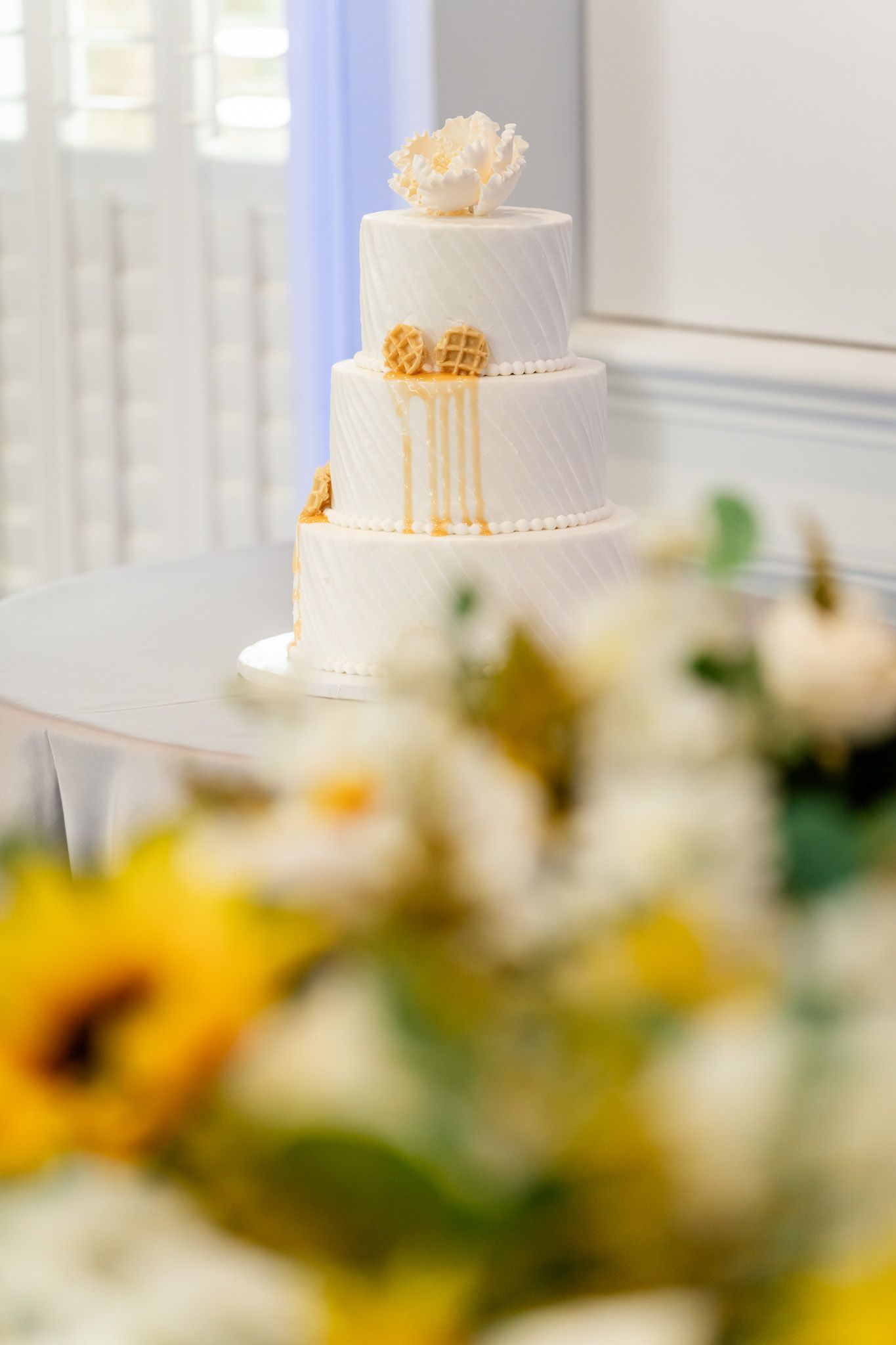 Elegant three-tier white wedding cake with gold drizzle and shell topper near sunflower bouquet.