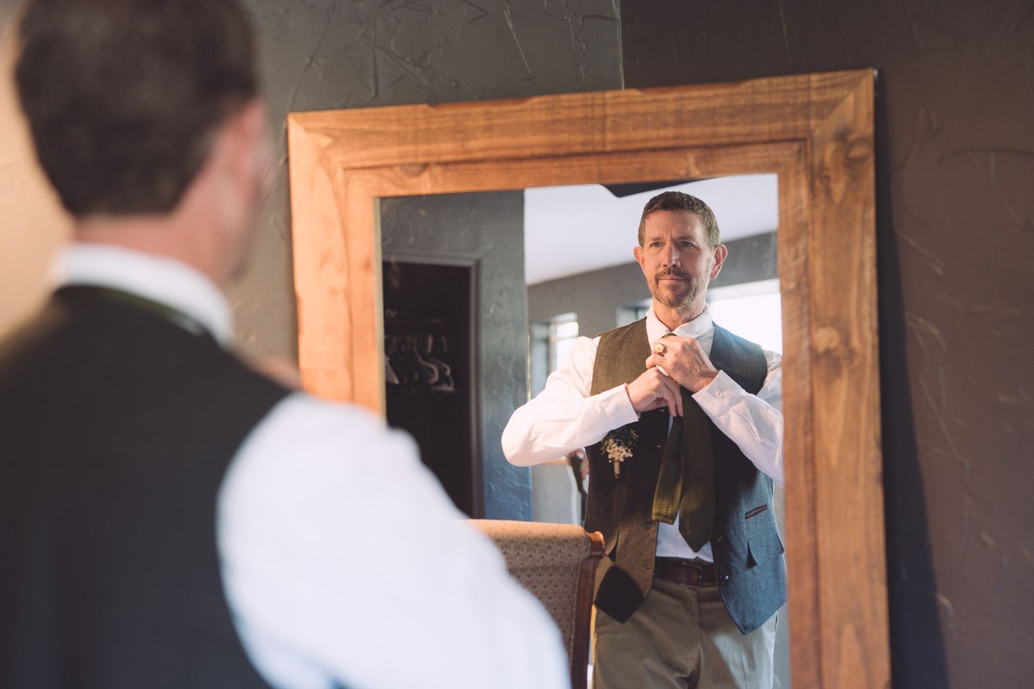Groom adjusts vest while checking appearance in rustic wooden framed mirror before wedding.