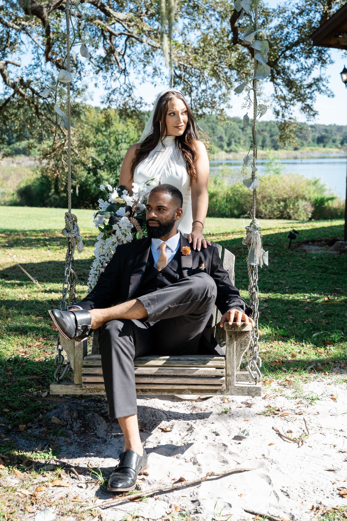 Bride stands behind seated groom on a rustic outdoor swing with lake views and lush greenery.