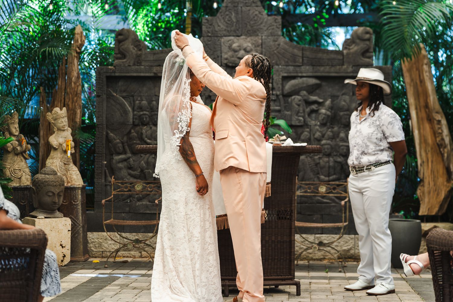 Bride in peach suit lifts veil of bride in lace gown at outdoor wedding ceremony.