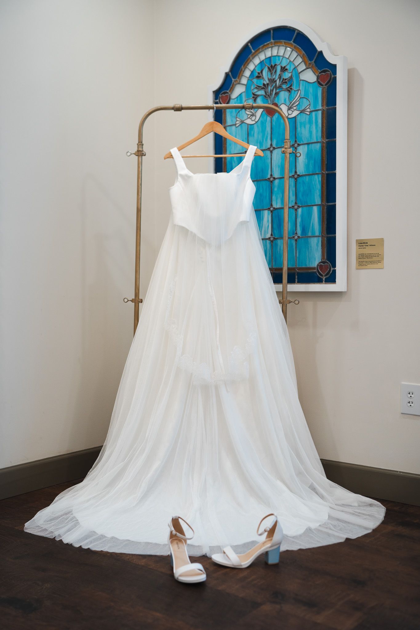 Elegant white wedding gown hanging on rack before stained glass window with heels below.