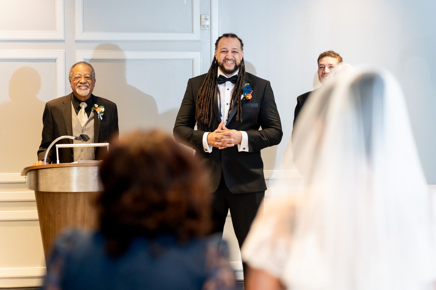 Groom with dreadlocks smiles joyfully as bride approaches during indoor wedding ceremony.
