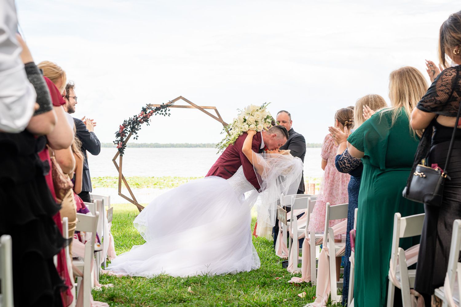Newlyweds share first kiss while guests celebrate along the aisle at outdoor lakeside wedding ceremony.