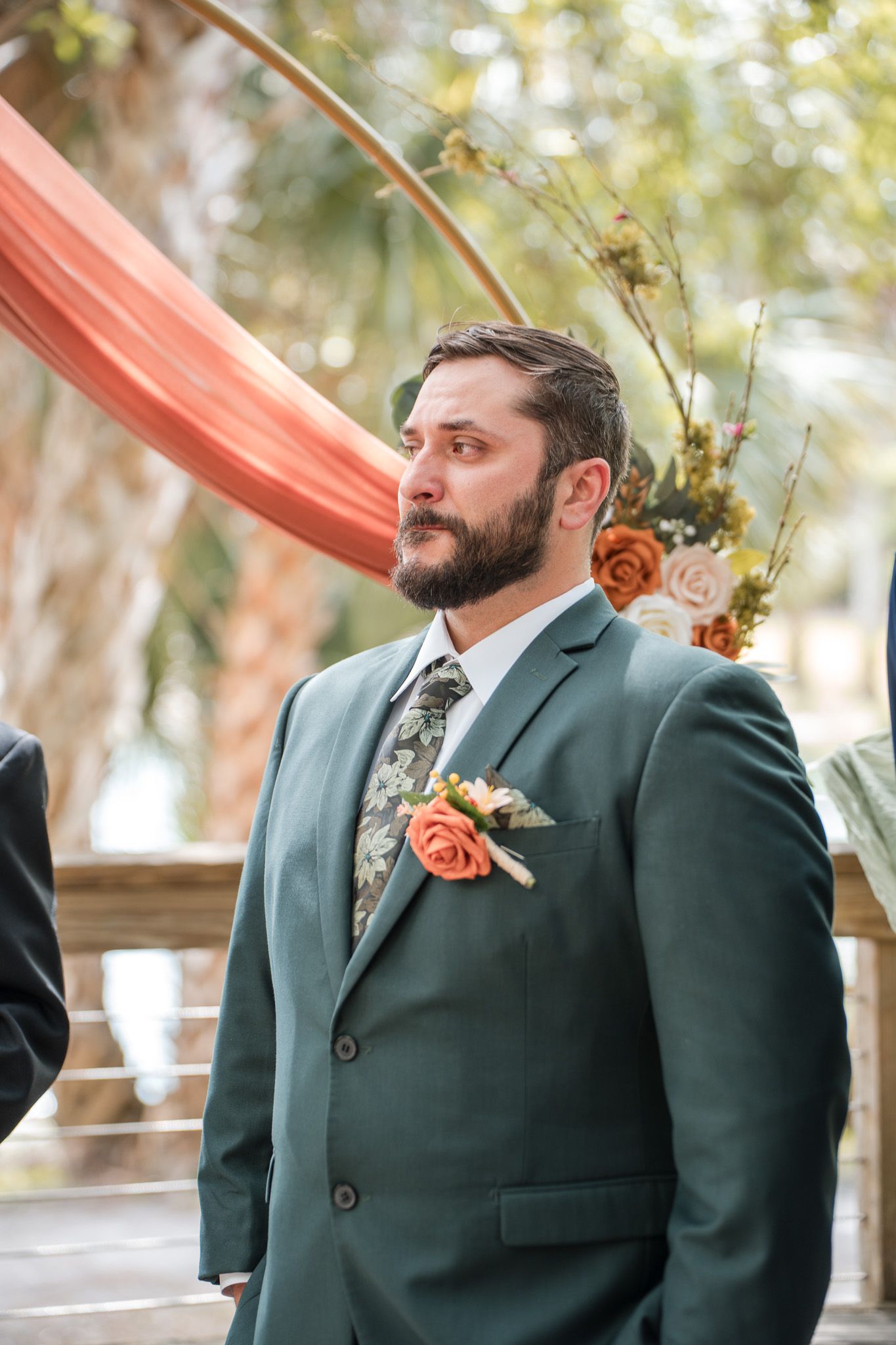 Groom in green suit with floral boutonniere stands at wedding altar decorated with coral draping and flowers.