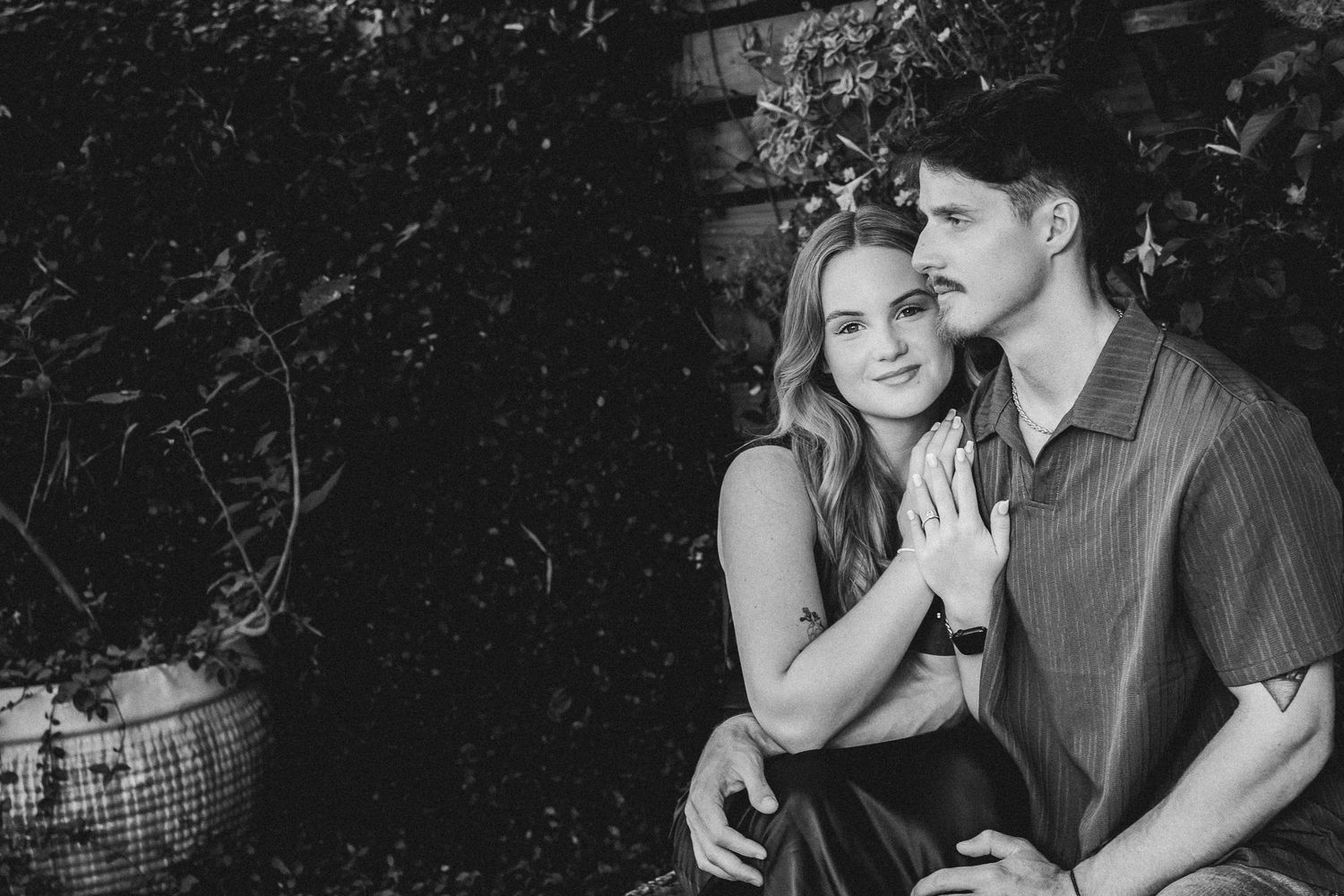 Black and white photo of a young couple sitting together outdoors, woman leaning on mans shoulder smiling.