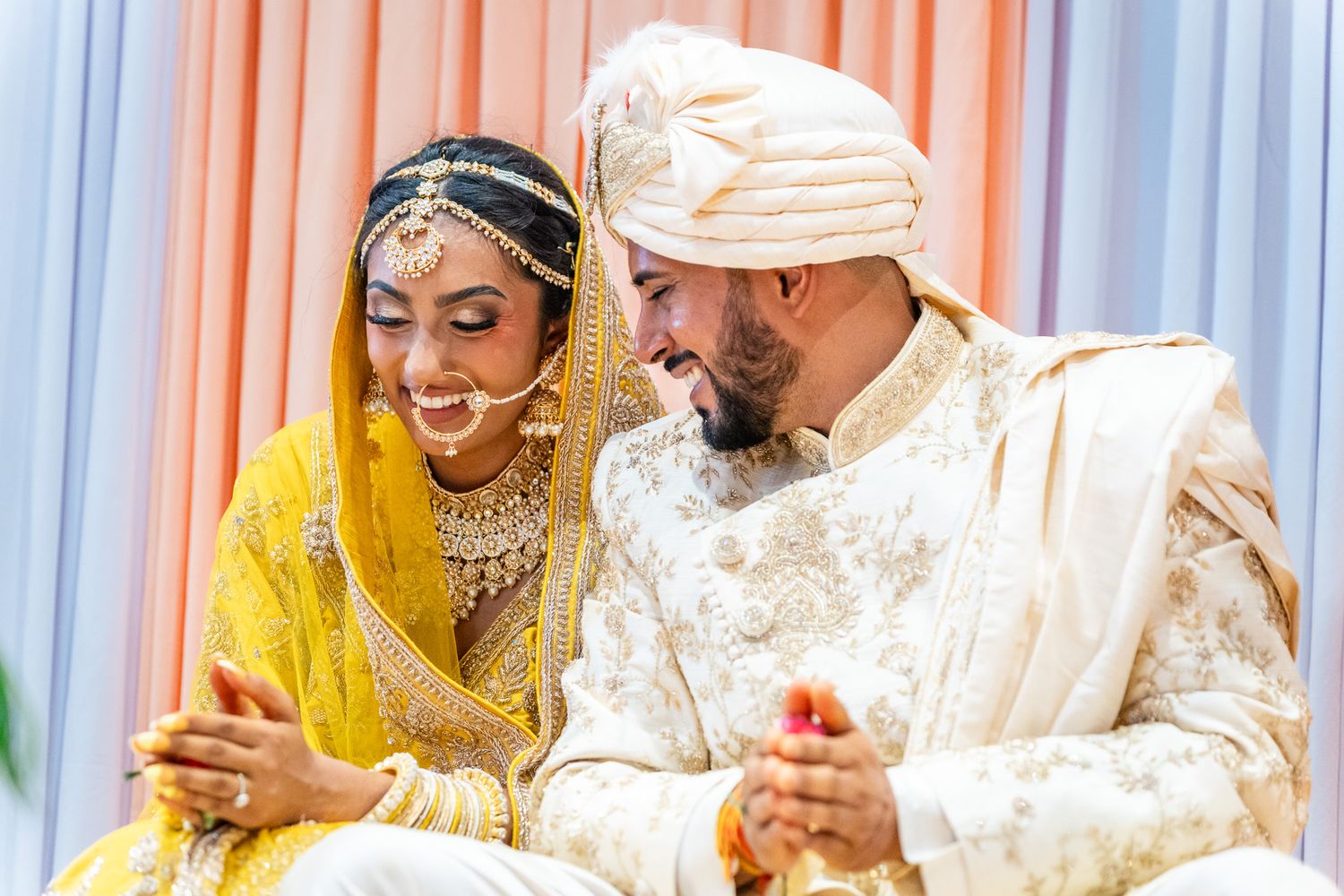 Indian bride and groom in traditional yellow and white wedding attire laugh together during ceremony.