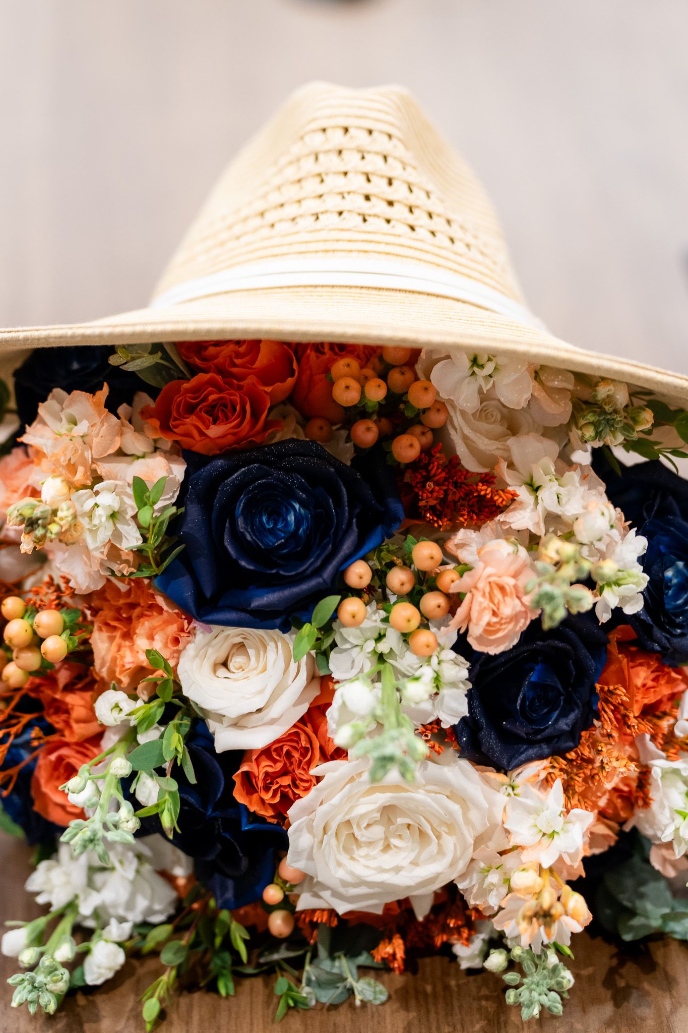 Vibrant wedding bouquet of navy blue, orange, and white roses with berries under a straw hat.