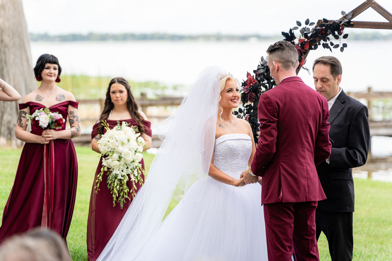 Bride in white gown holds bouquet facing groom in burgundy suit at outdoor wedding ceremony with bridesmaids.