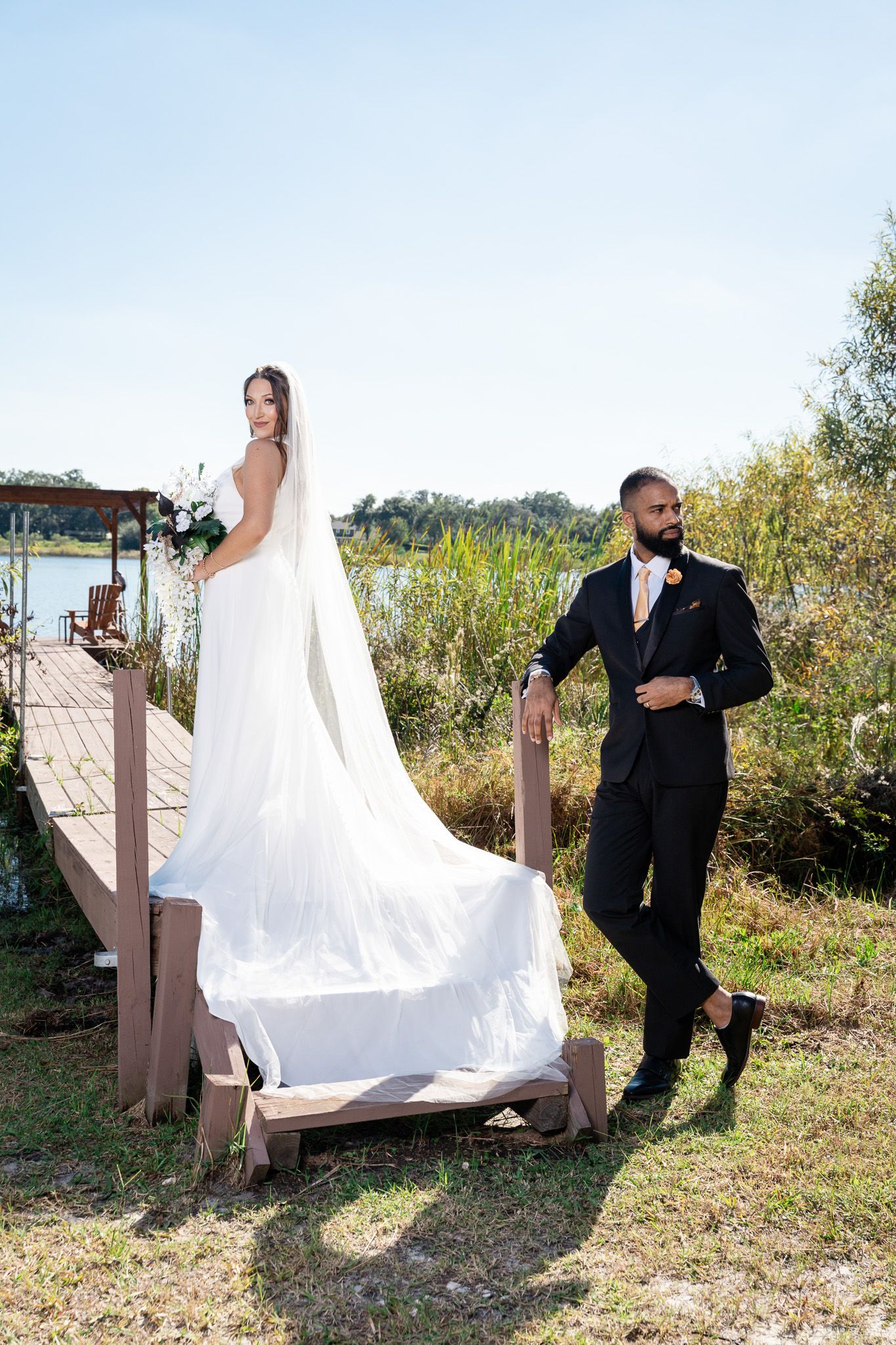 Bride in white gown with long veil stands on dock as groom in black suit adjusts jacket near lakeside.