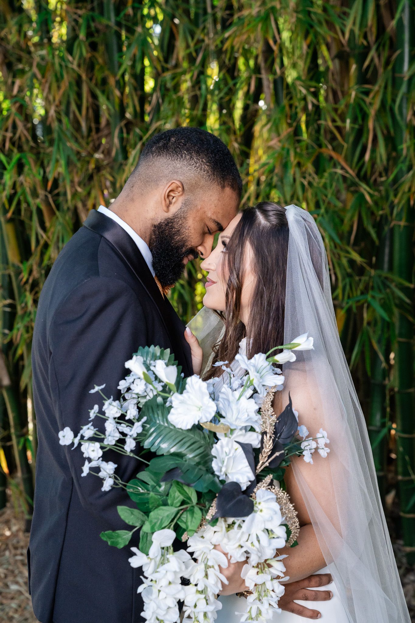 Bride and groom touch foreheads tenderly as she holds white floral bouquet before bamboo trees.
