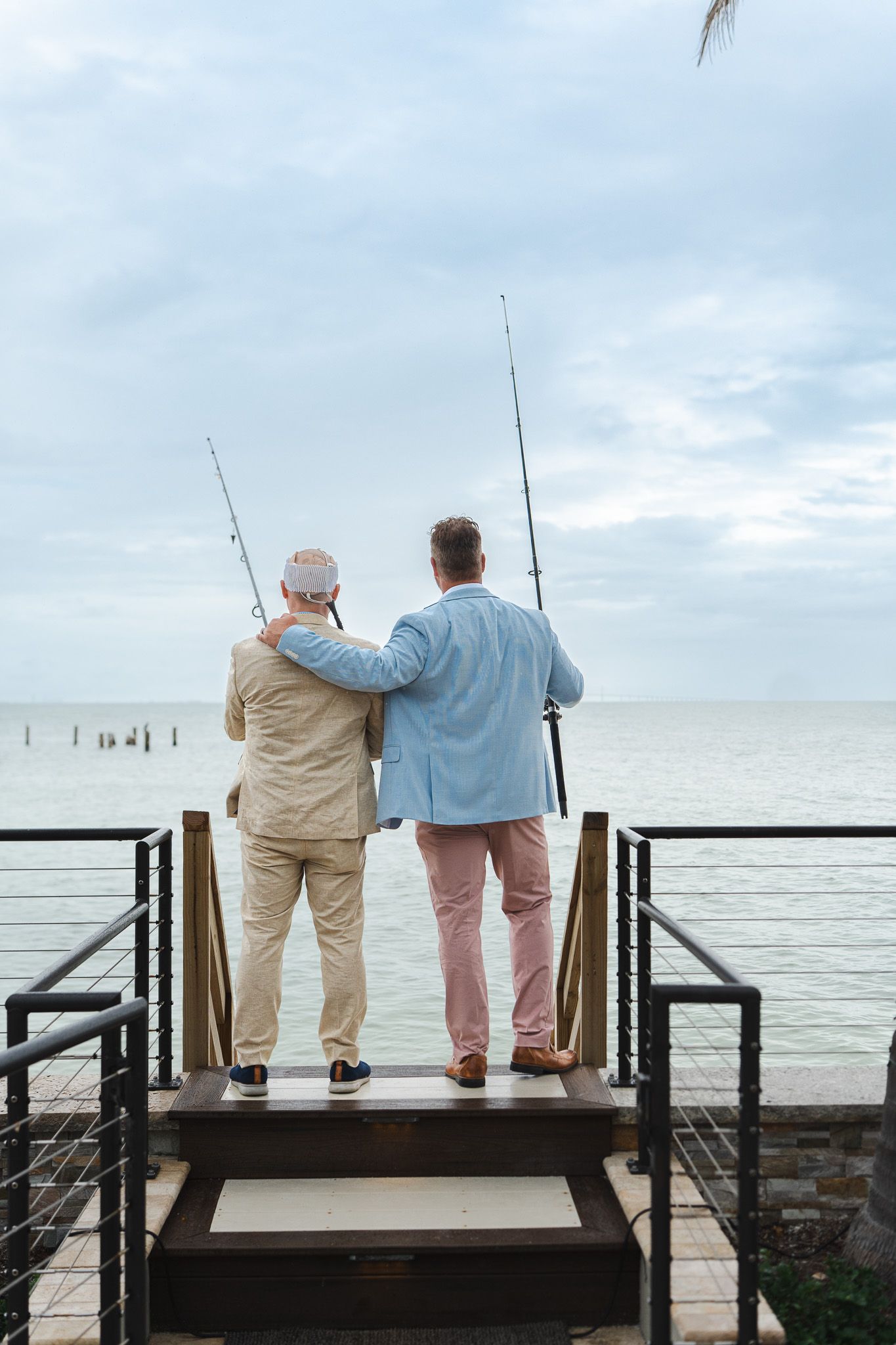 Two men stand arm in arm holding fishing rods on a waterfront dock overlooking the calm sea.