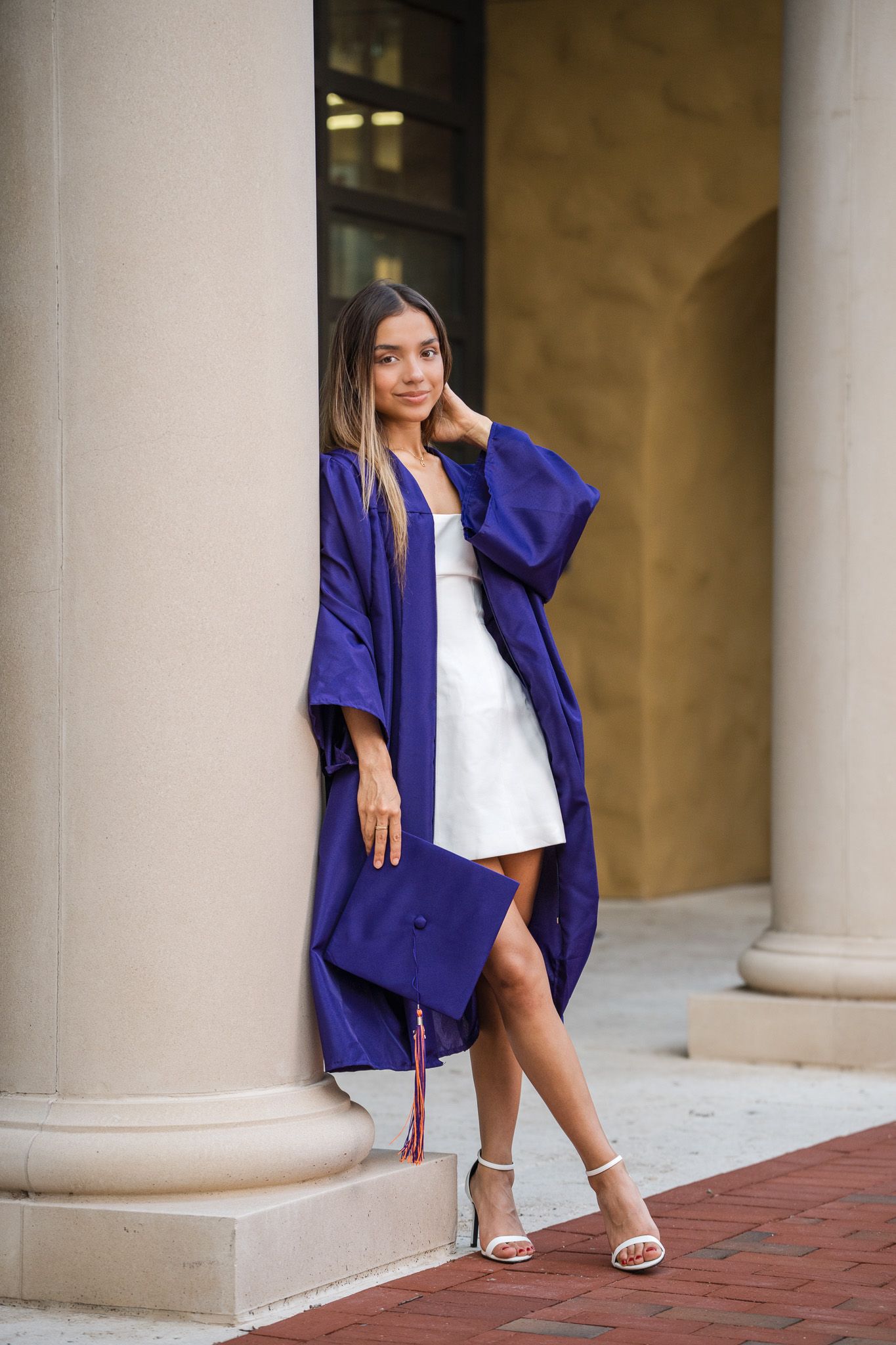 Female graduate in blue gown leans against column holding cap on a classic university building.