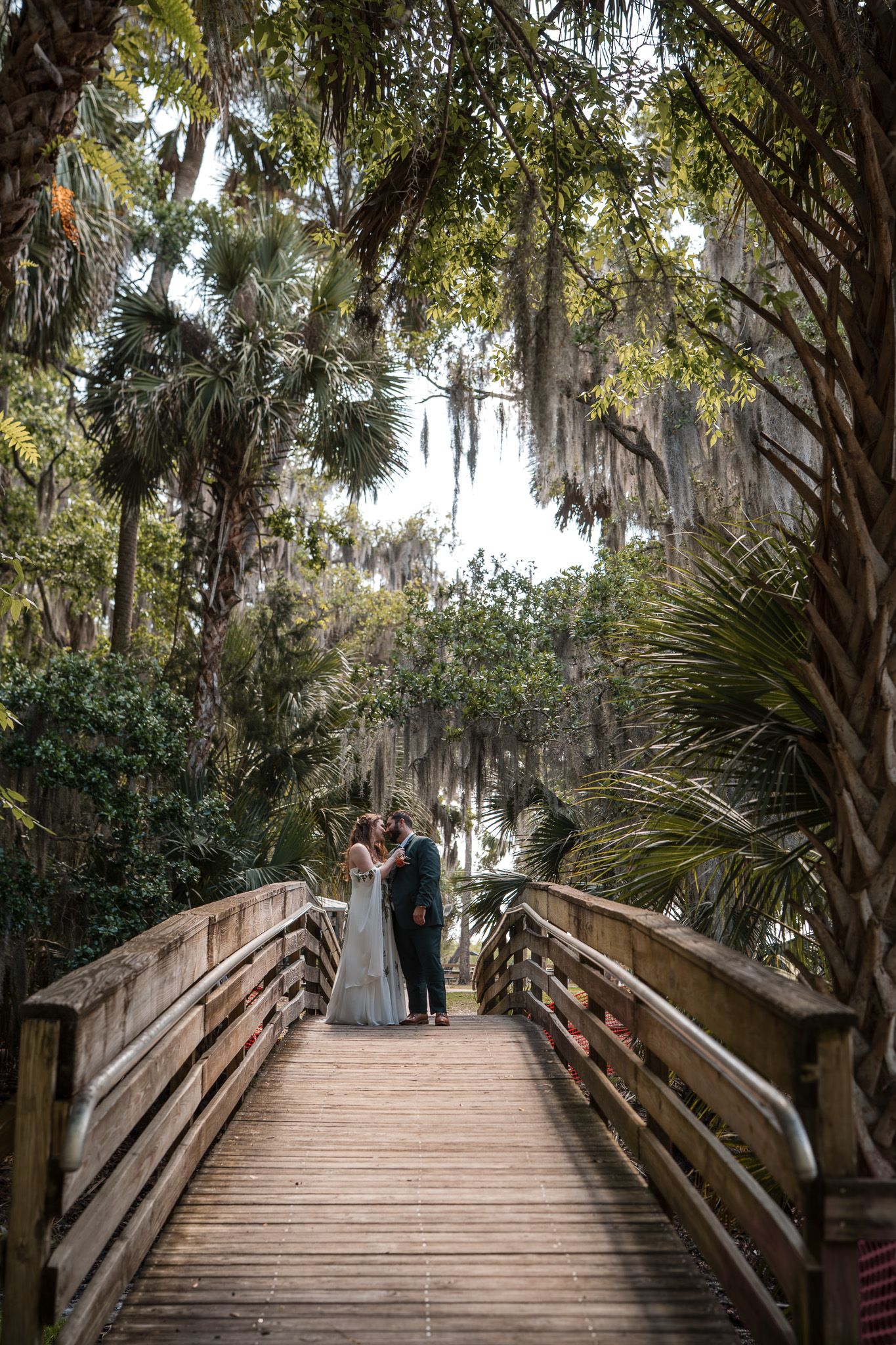 Bride and groom share romantic moment on wooden bridge canopied by palm trees and Spanish moss.