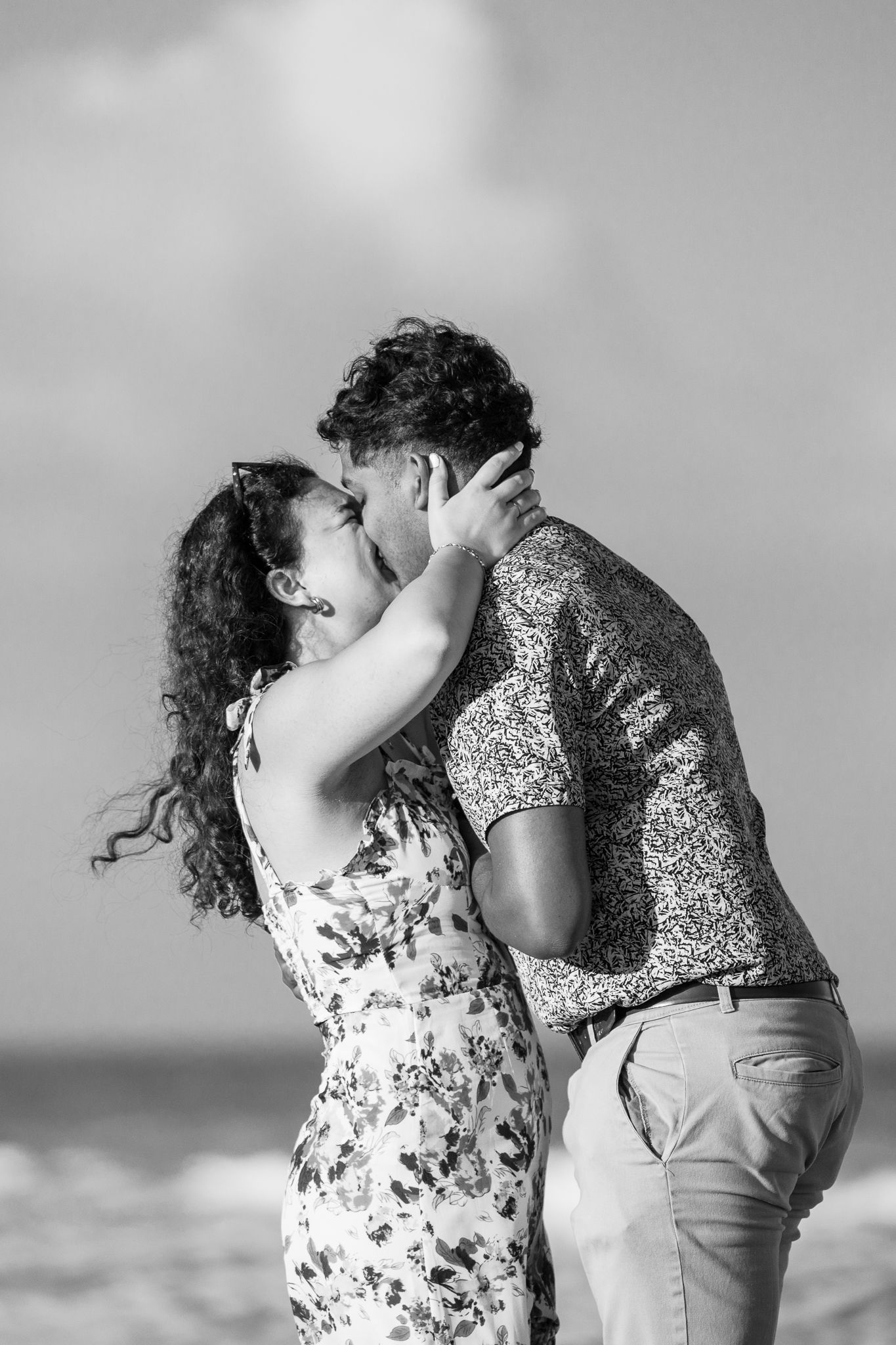 Black and white photo of a couple kissing romantically on a beach, woman in floral dress, man in patterned shirt.
