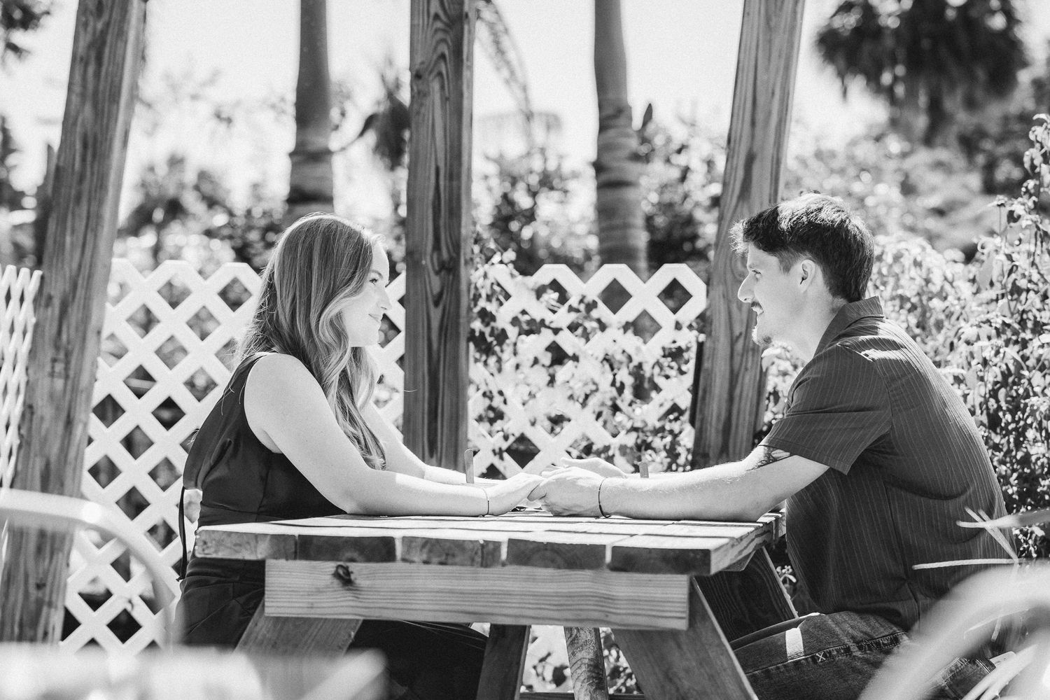 Black and white photo of a couple holding hands at a picnic table outdoors, surrounded by lush tropical foliage.
