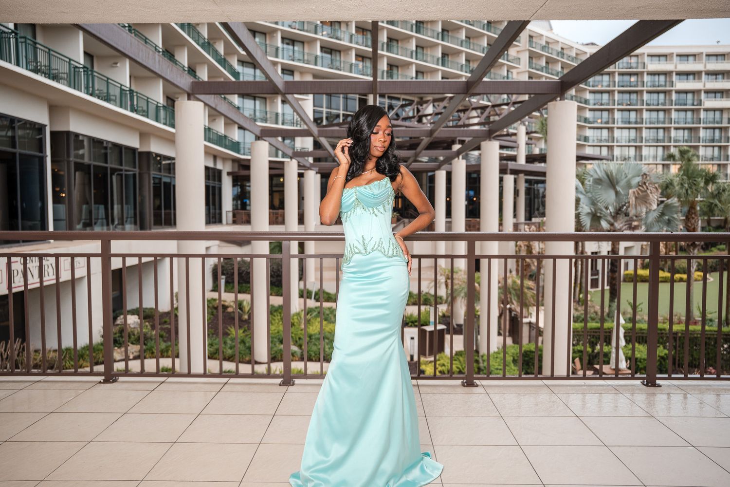 Elegant woman in mint green mermaid dress posing on outdoor hotel balcony with courtyard view.