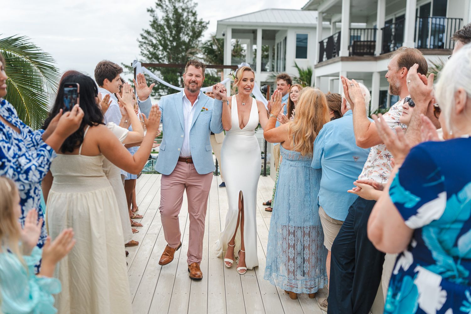 Newlyweds walk down aisle as guests cheer and applaud at sunny outdoor beach wedding reception.
