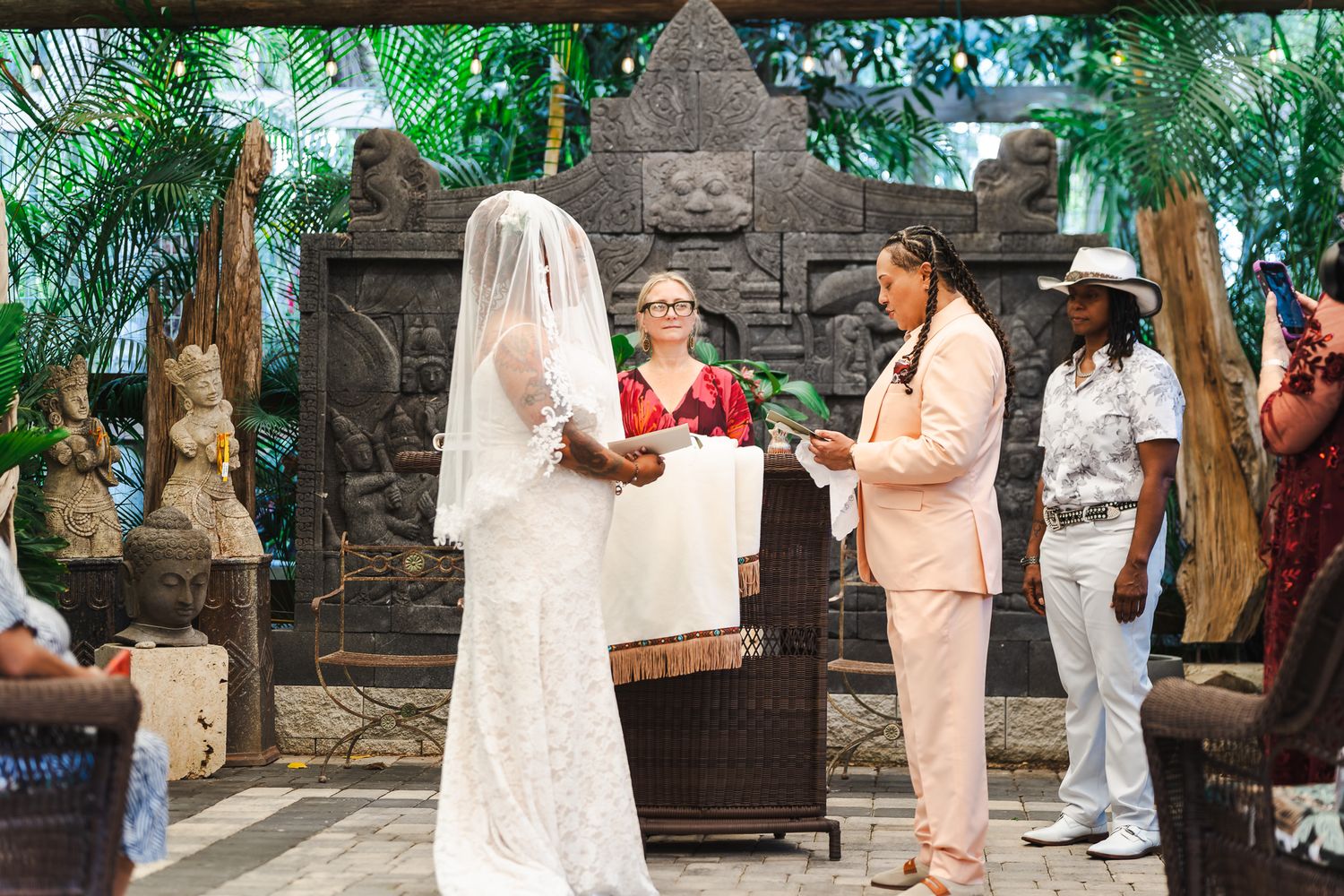 Same-sex wedding ceremony outdoors with two brides and officiant surrounded by tropical greenery.