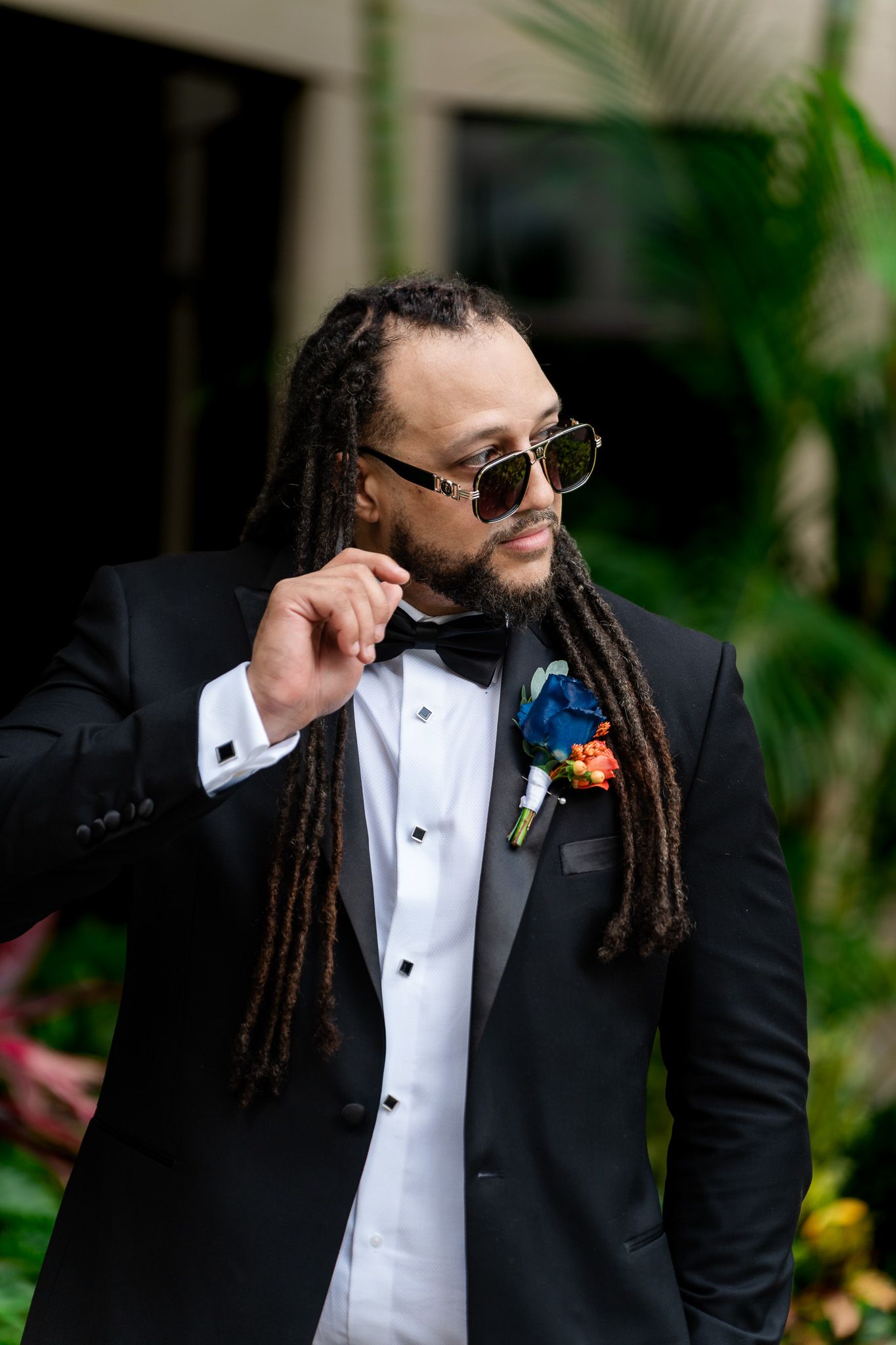 Stylish groom in black tuxedo with dreadlocks, sunglasses, and colorful boutonniere poses outdoors.