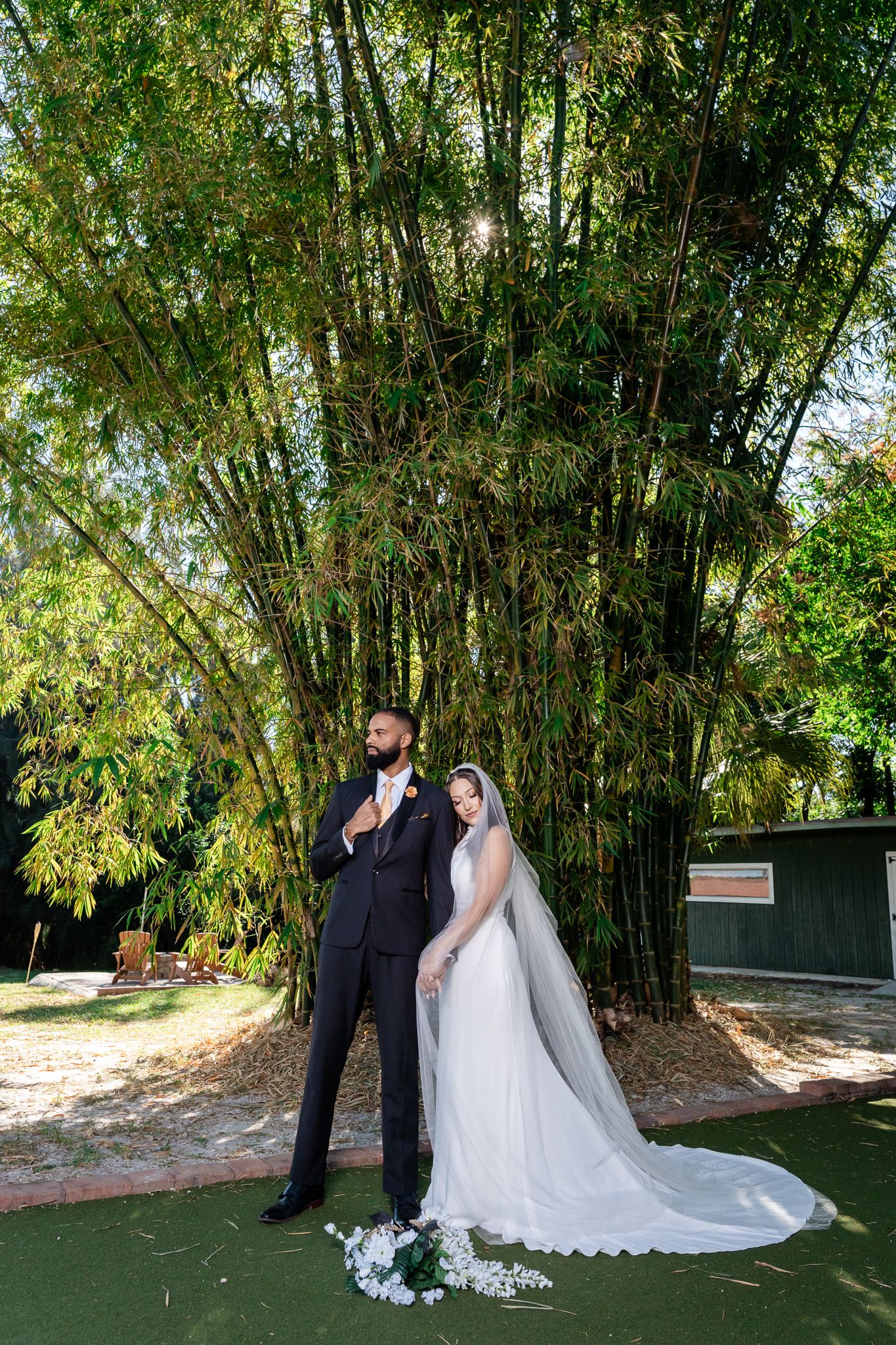 Bride in white gown with long veil stands with groom in black suit before tall bamboo trees.