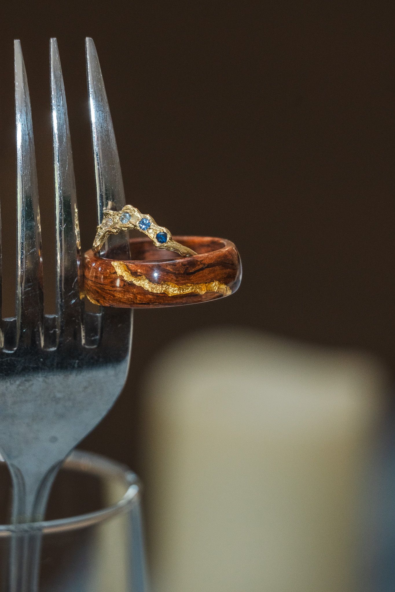 Unique wooden and gold wedding rings balanced on a silver fork with candle bokeh background.