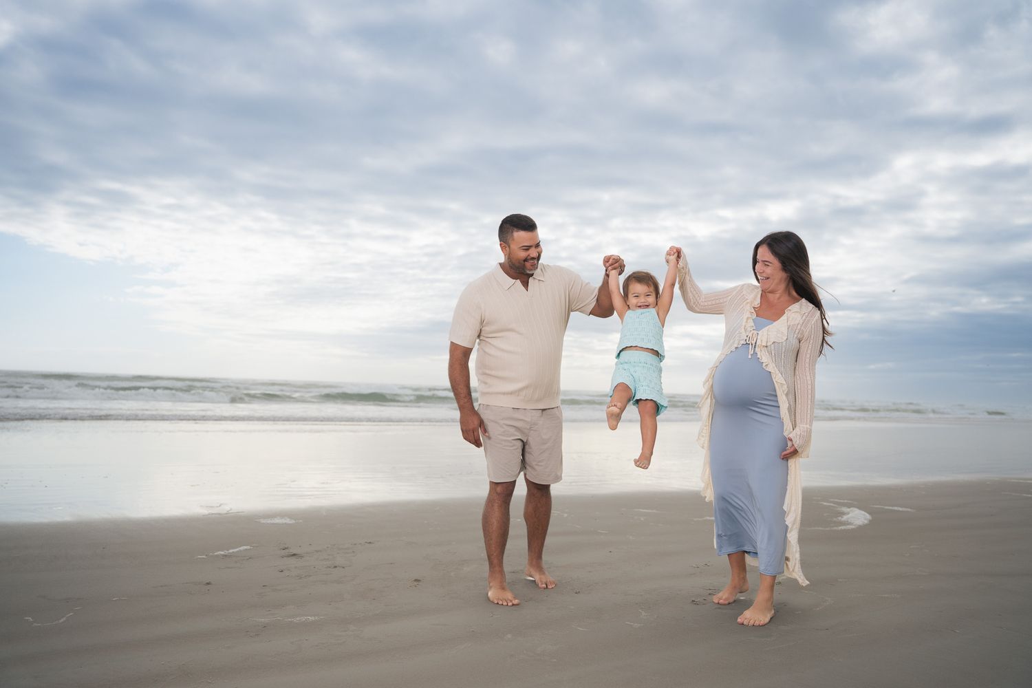 Expecting family swings toddler between parents on a serene beach with cloudy skies in the background.