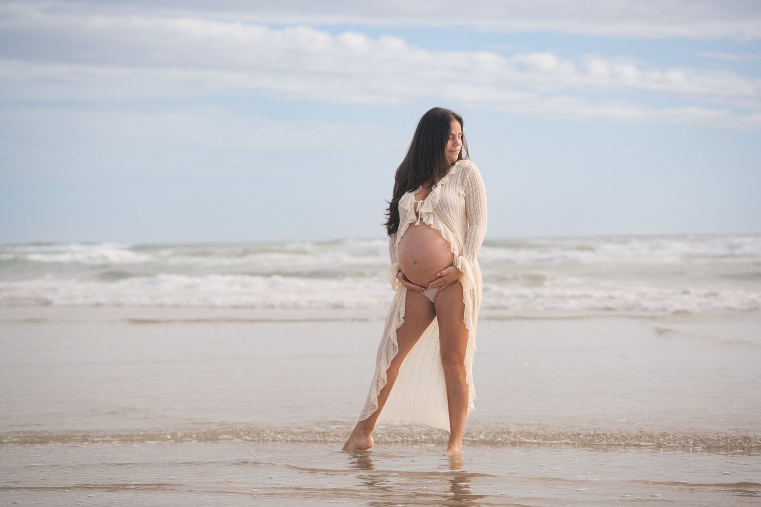 Pregnant woman in sheer lace cover-up walking along ocean shoreline at beach.