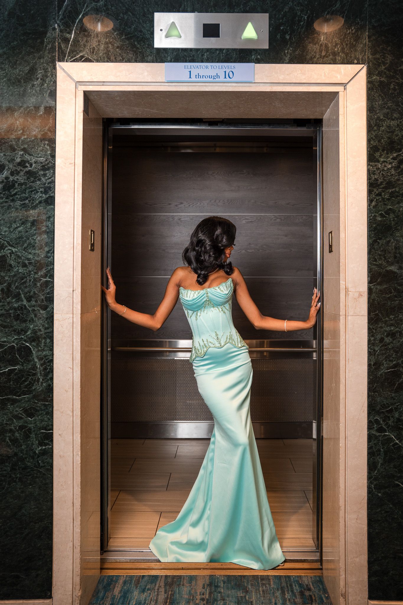 Woman in elegant teal mermaid gown posing inside a luxury elevator with marble walls.