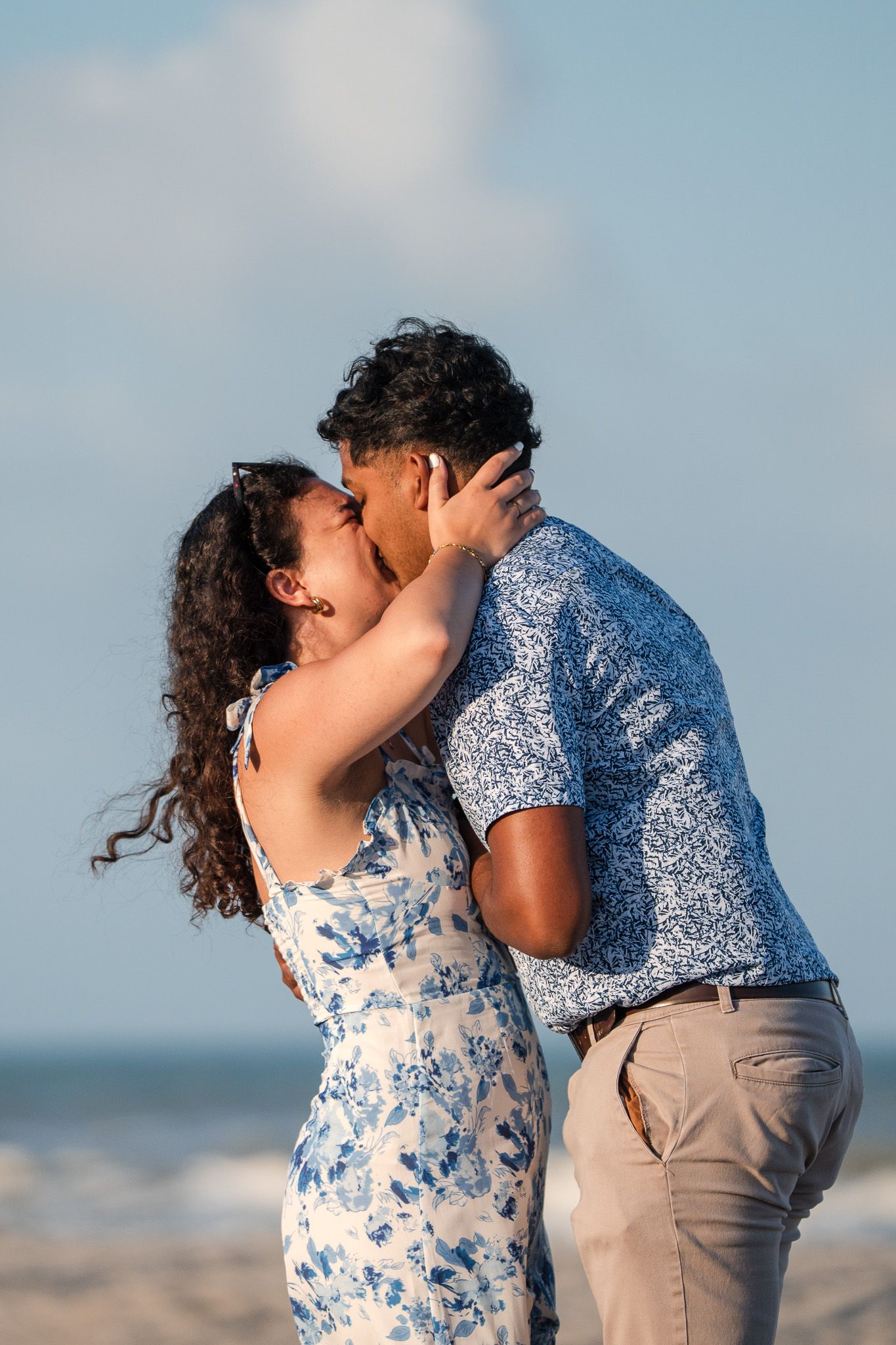 Couple sharing a romantic kiss on the beach, dressed in matching blue and white outfits with ocean waves behind them.
