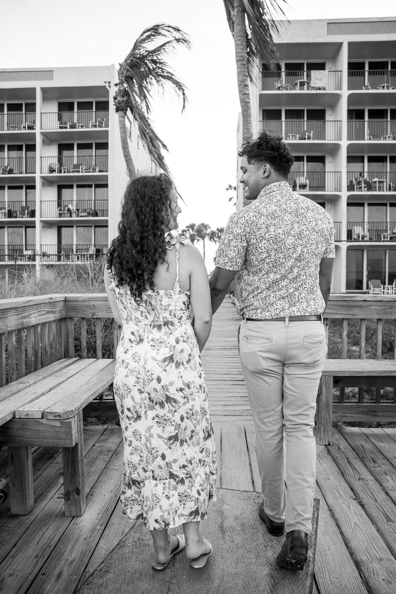 Black and white photo of a couple holding hands walking on a wooden boardwalk toward a beachside hotel with palm trees.