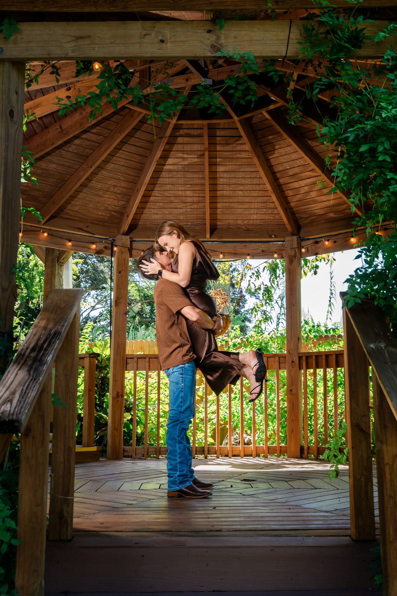 Couple sharing a romantic embrace in a wooden gazebo surrounded by lush green vines and string lights.