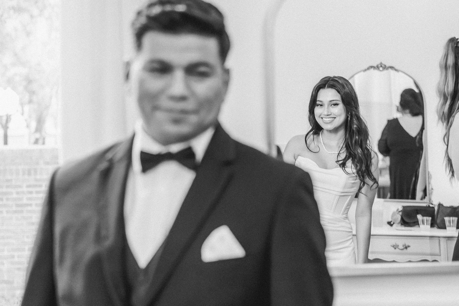 Groom in black tuxedo stands in foreground as bride smiles in mirror reflection behind him.