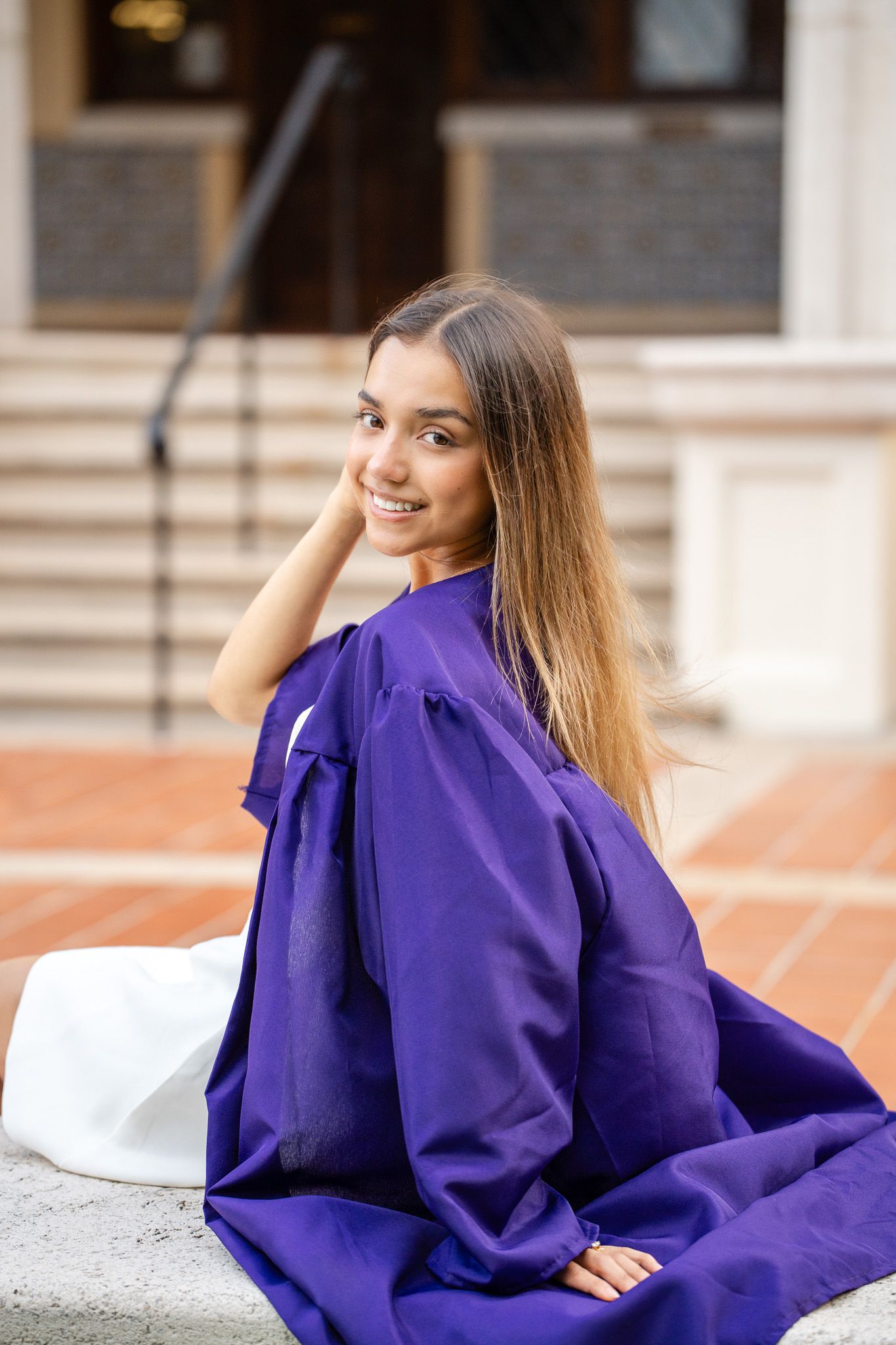 Smiling young woman in purple graduation gown sitting on steps, posing for senior portrait at university.