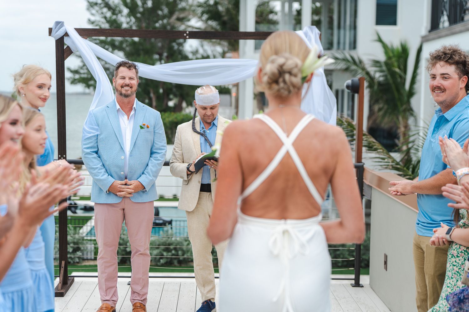 Bride in backless white gown walks toward smiling groom at outdoor wedding ceremony altar.