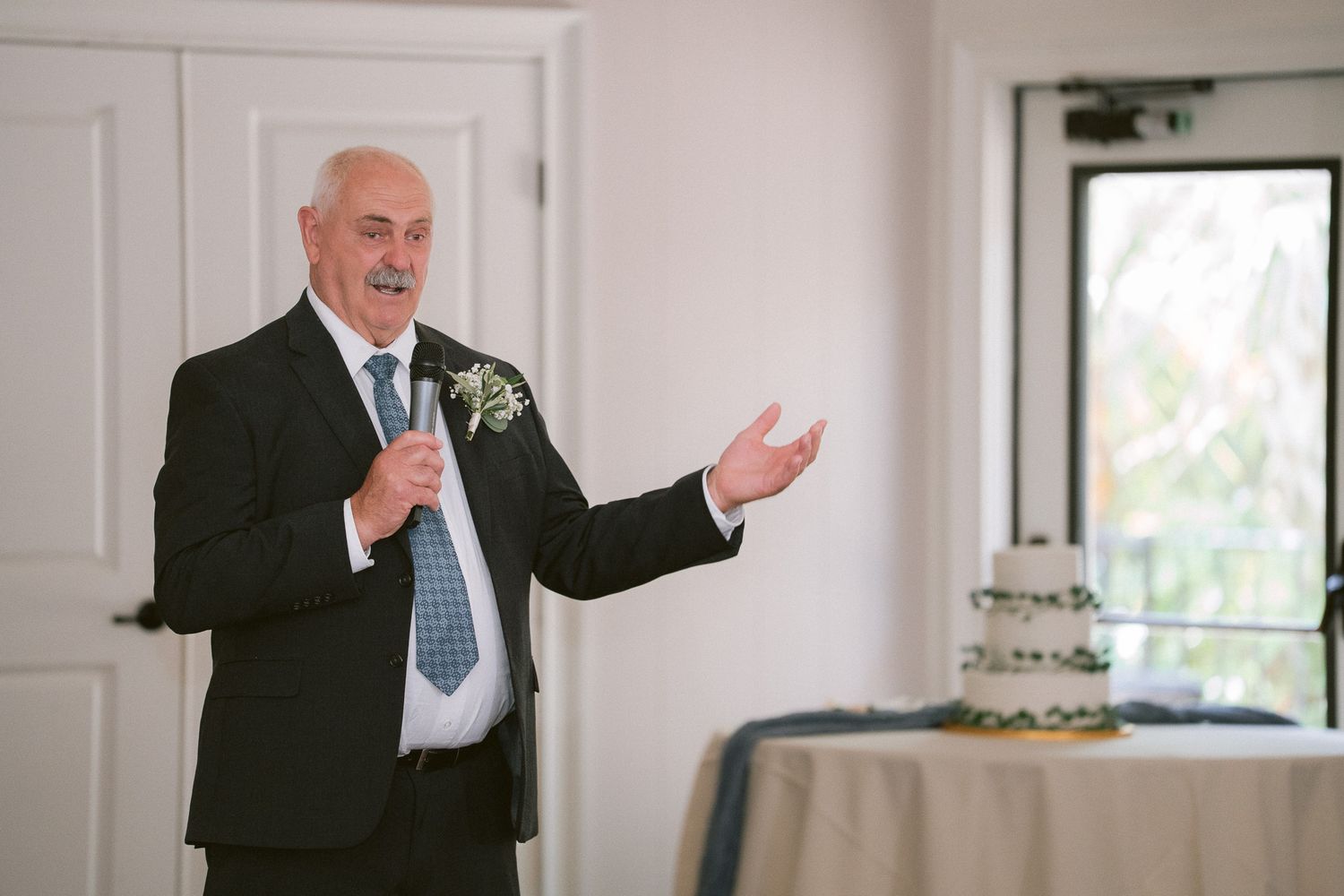 Father of the bride delivers heartfelt wedding speech beside tiered cake.