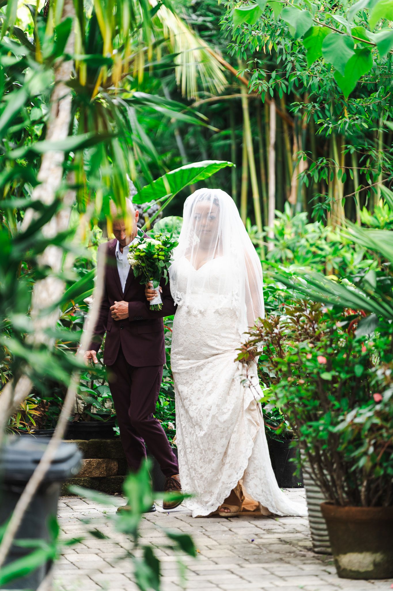 Bride in lace gown and veil walking with groom in burgundy suit through a lush tropical garden.
