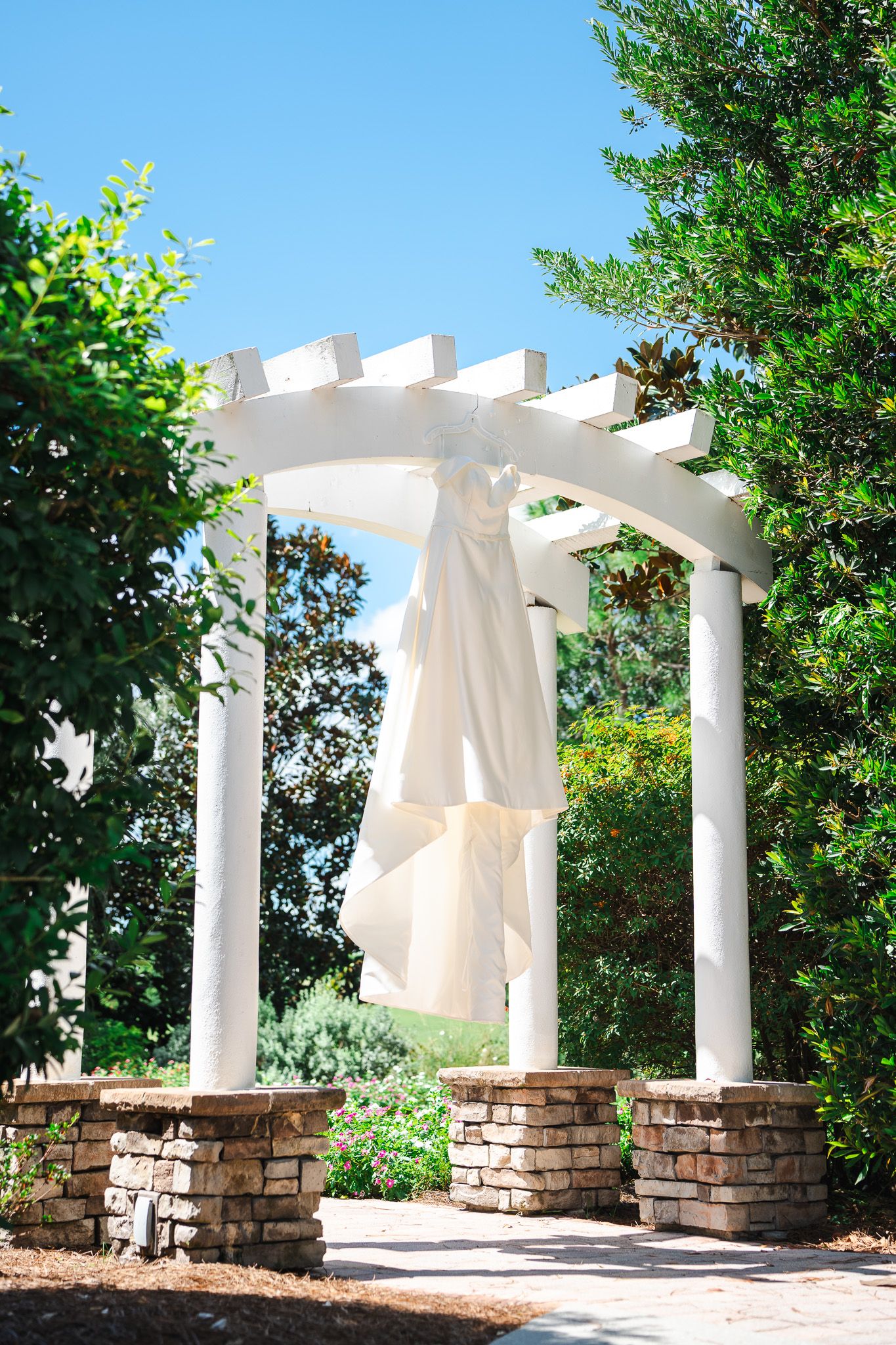 White wedding dress hanging on elegant pergola arch surrounded by lush green trees and blue sky.