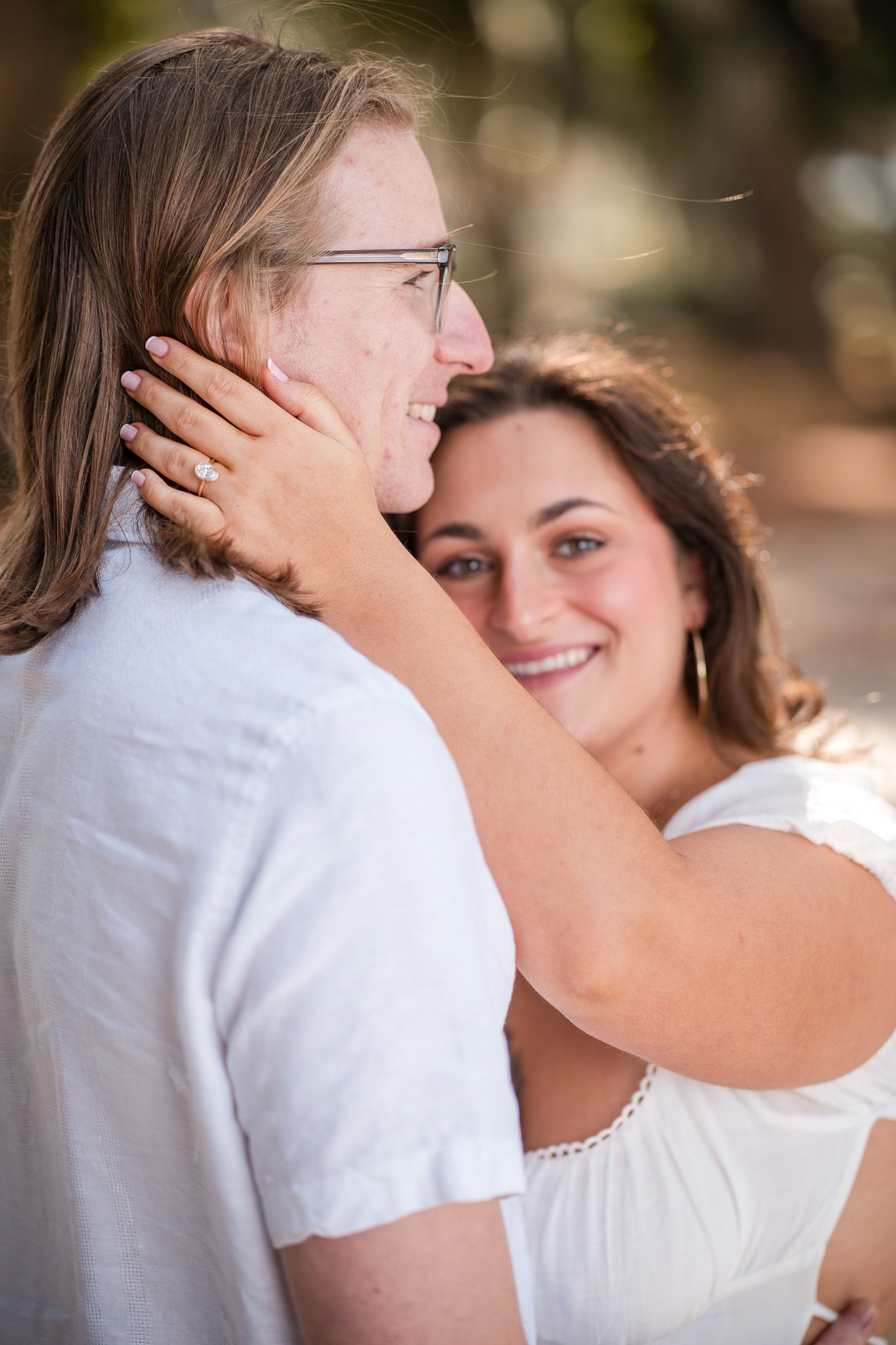 Woman smiles warmly while gently cradling mans face showing engagement ring during outdoor photo session.