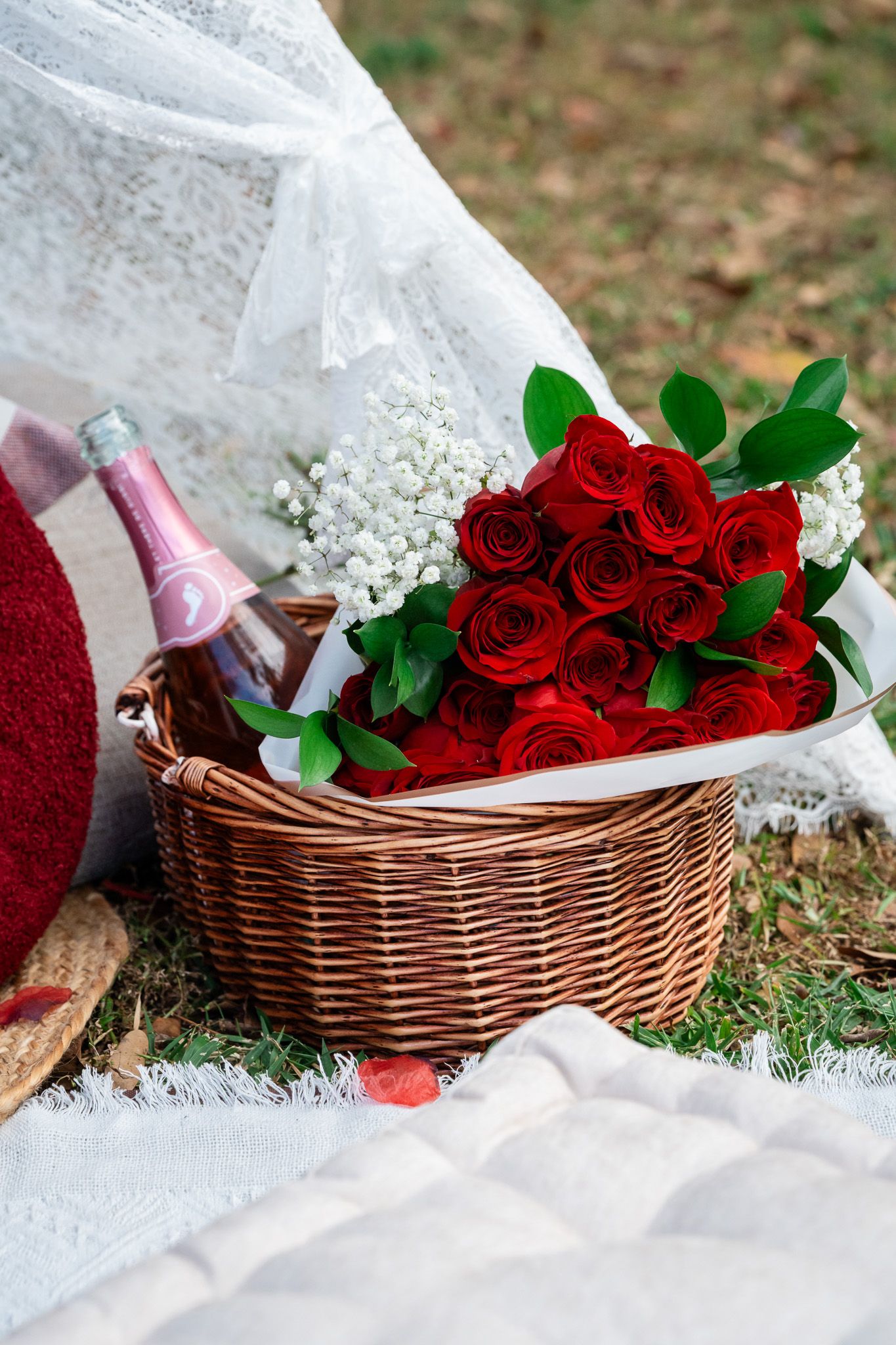 Wicker basket with red roses bouquet and pink champagne bottle on white blanket for romantic outdoor picnic.