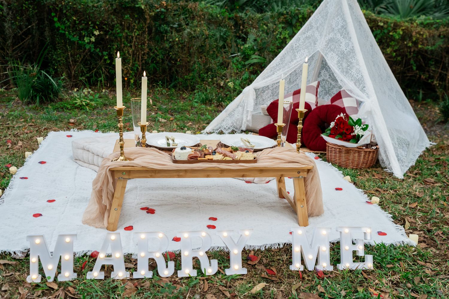 Romantic outdoor proposal setup featuring a lace teepee, candles, roses, and Marry Me sign.