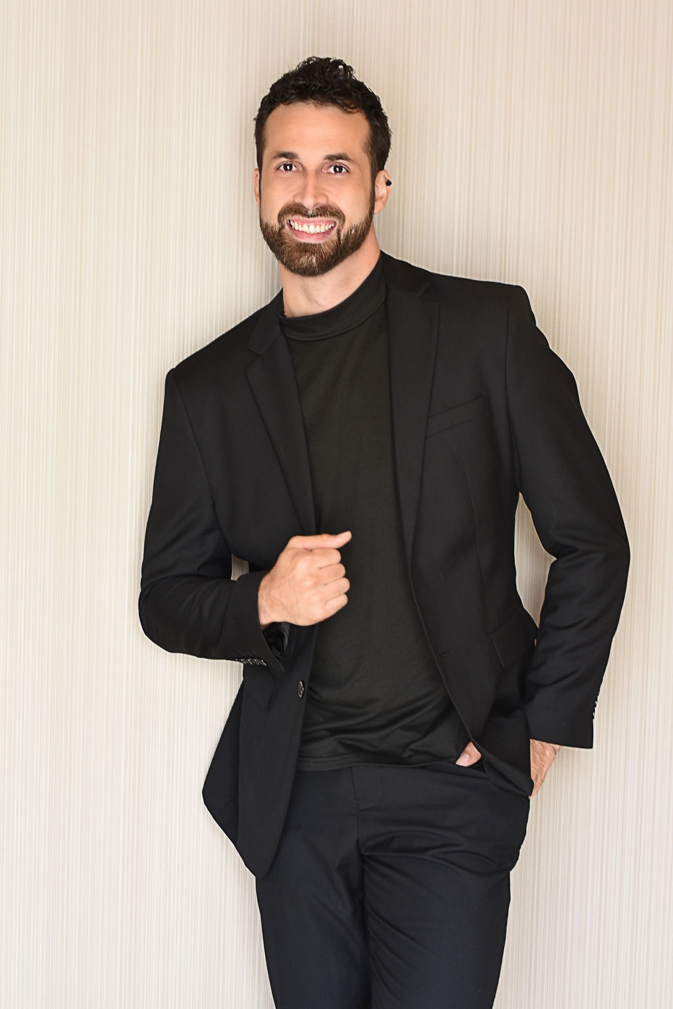 Smiling man with beard wearing all-black suit and turtleneck, posing confidently against a light textured wall.